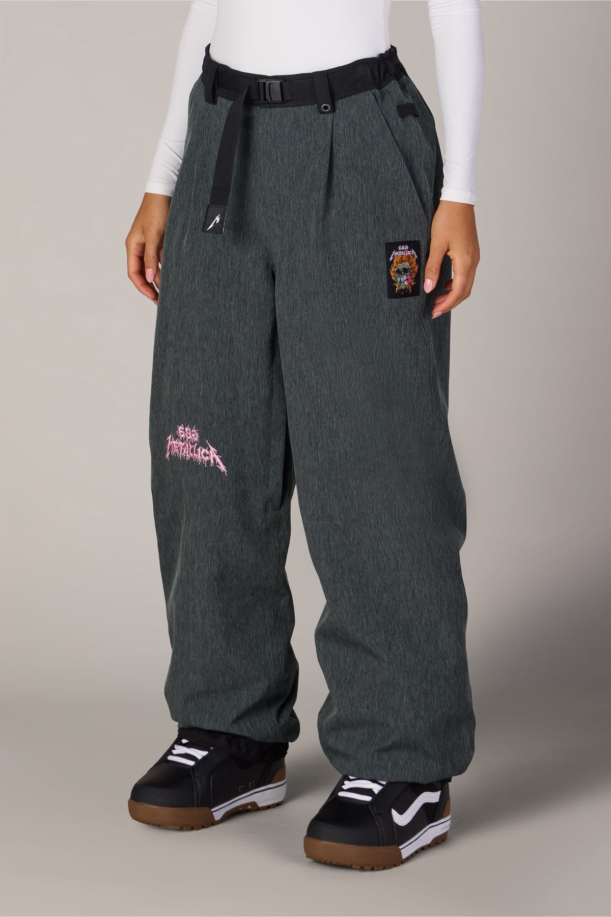 686 Women’s Metallica Burning Skull Shell Pant in dark grey, featuring a waterproof woven design, a burning skull graphic on the thigh, a small hip patch, and a black webbed belt at the waist.