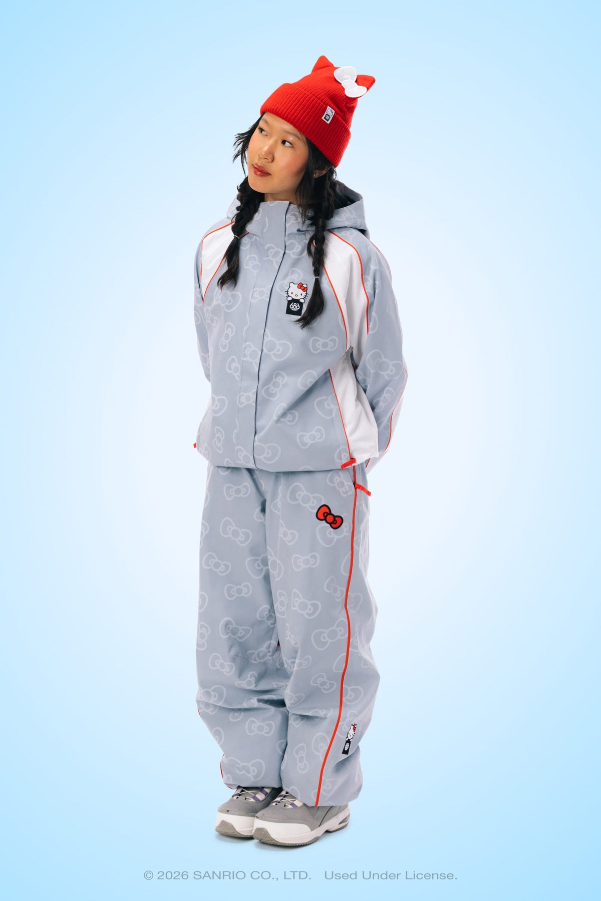A woman wearing the 686 Women’s Hello Kitty® Lil Bow Shell Pant and a red knit hat with a bow stands against a blue background, looking to the side with her hands in her pockets.