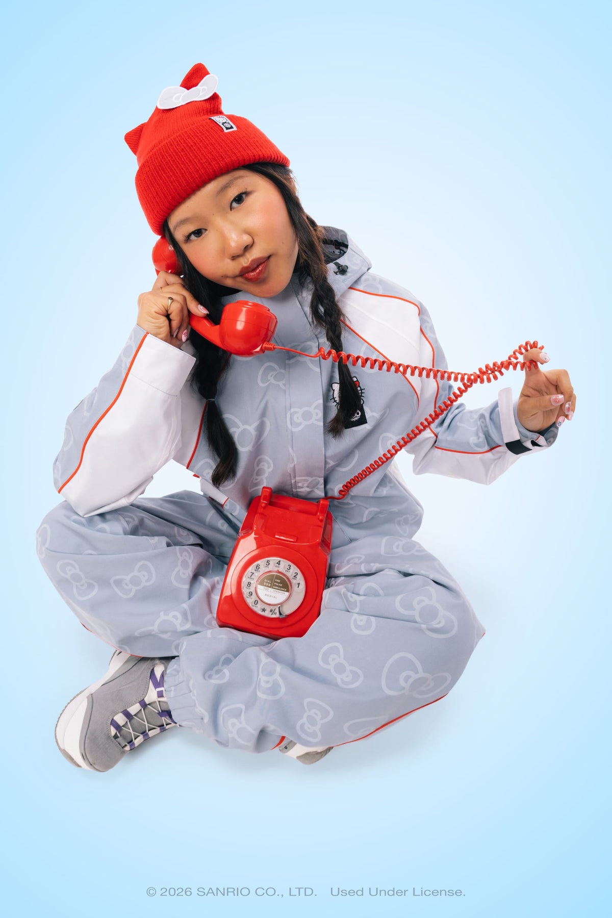A person in a red beanie sits cross-legged, holding a red retro rotary phone, while wearing the 686 Women’s Hello Kitty® Lil Bow Shell Pant by 686. The background is light blue.