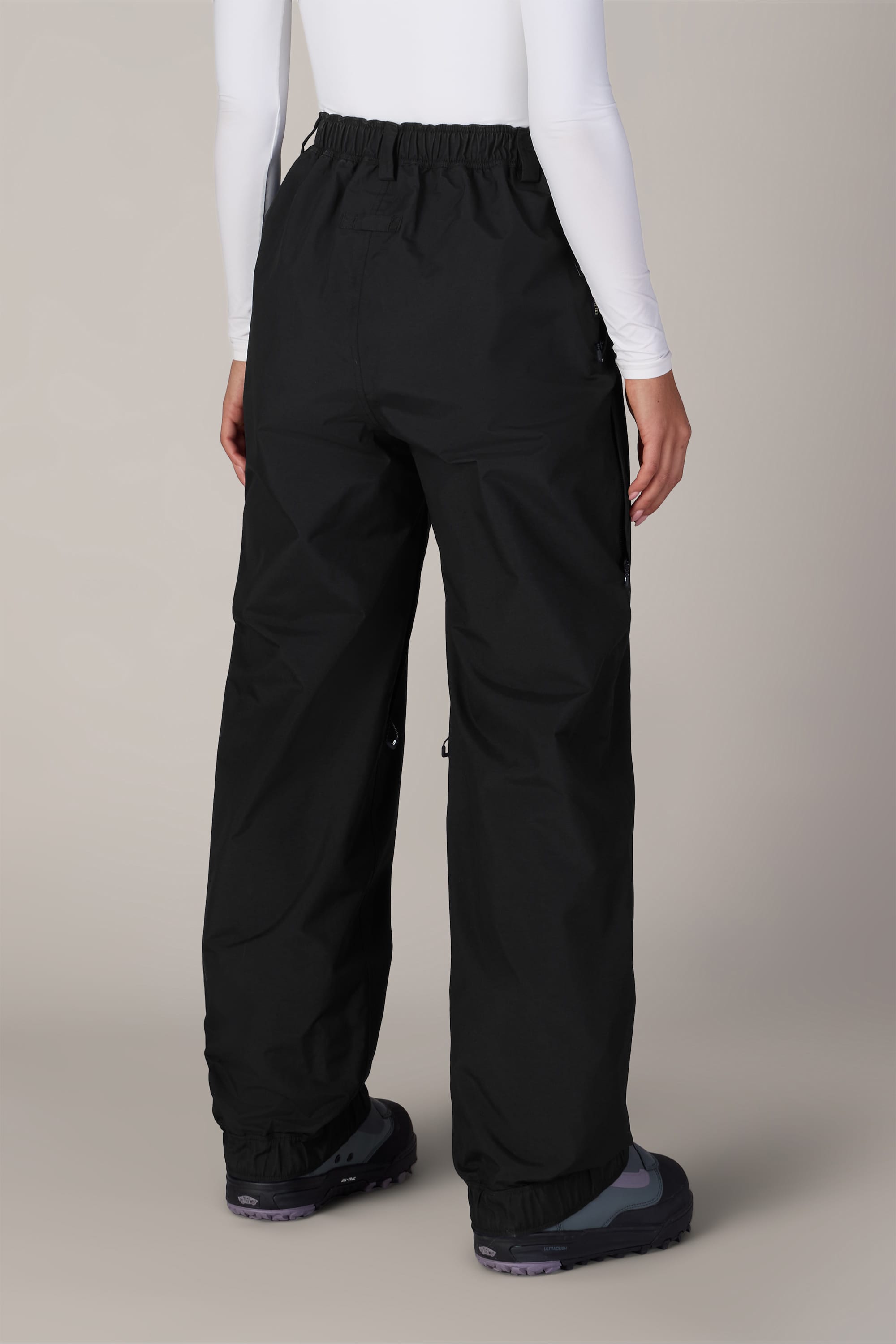 A person is seen from behind against a plain light background, wearing the 686 Women’s GORE-TEX® Outline™ Pant by 686—black, loose-fitting, waterproof nylon pants—paired with a long-sleeve white top and dark sneakers.