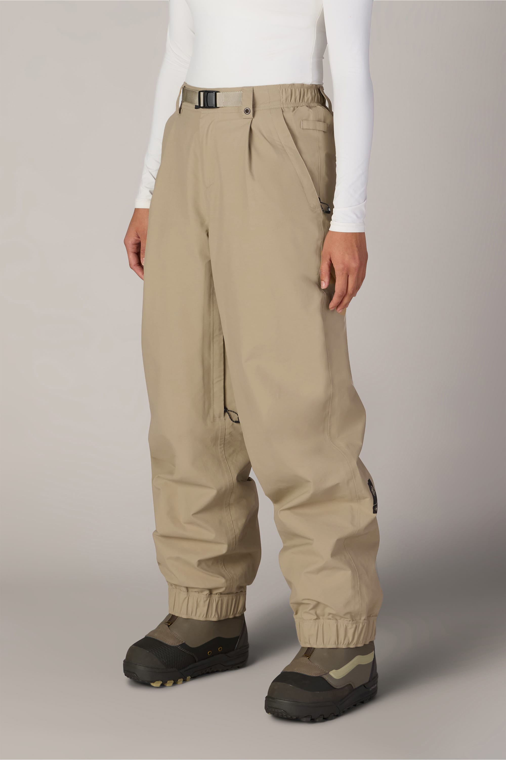 A person wearing the 686 Women’s GORE-TEX® Outline™ Pant—loose tan with elastic cuffs—paired with a white long-sleeve top and black-and-olive boots stands against a plain light background.