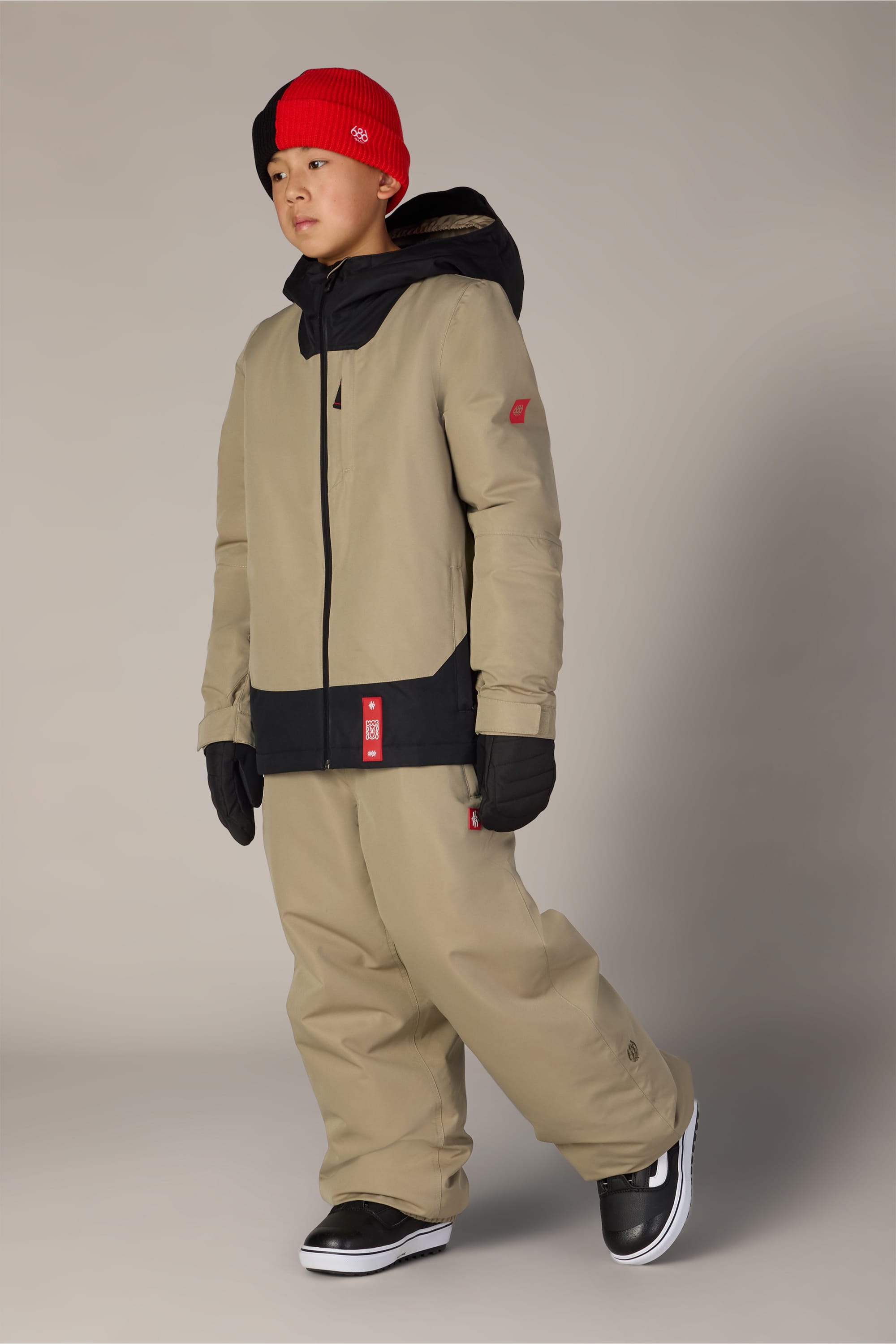 A person wearing the 686 Boys’ Gateway™ Insulated Jacket in tan, paired with matching pants, black gloves, black sneakers, and a red and black beanie stands against a plain light background.