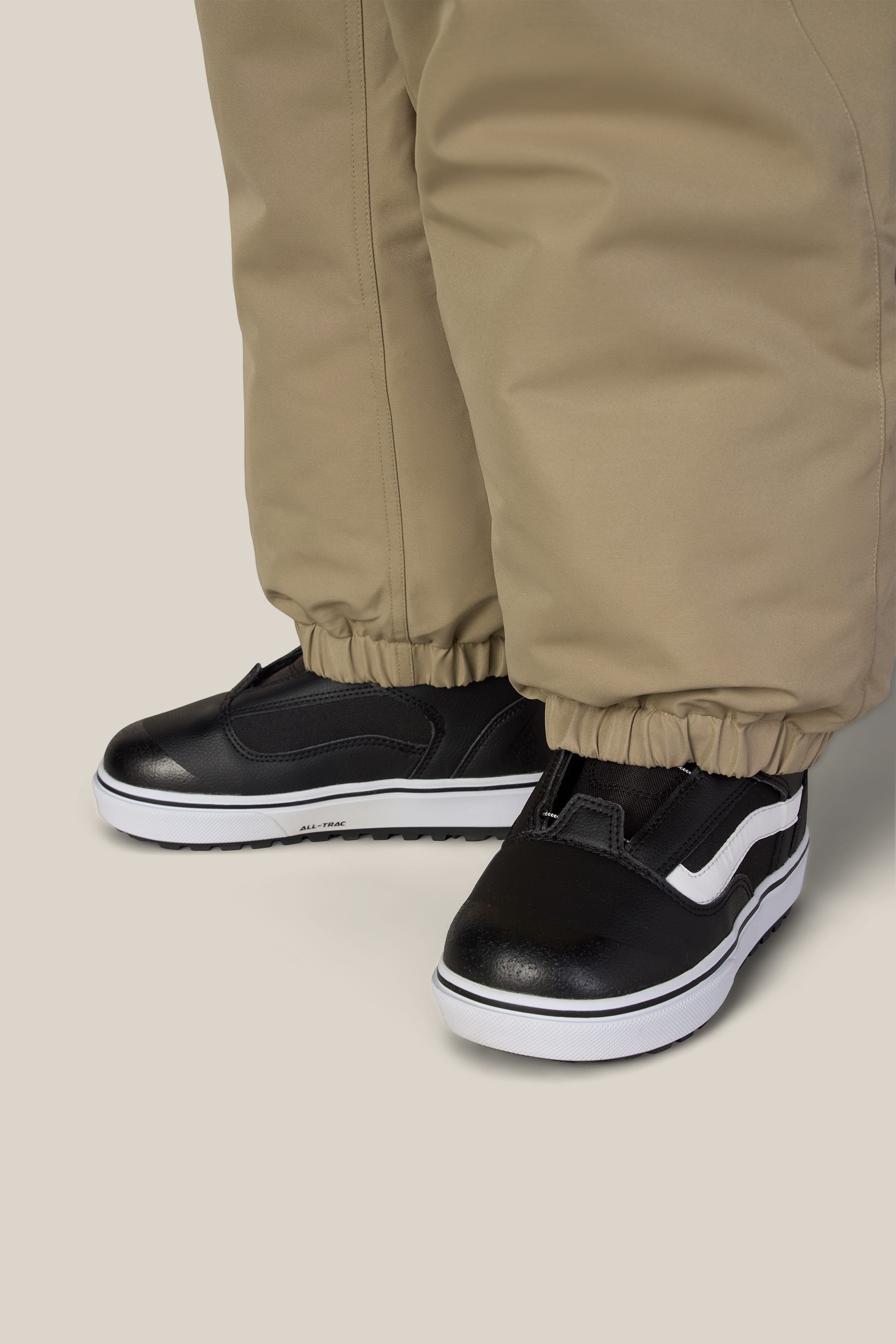 A child wearing the 686 Boys’ 686 Dojo® Insulated Pant in tan, featuring elastic ankles, paired with black slip-on sneakers with a white sole and side stripe, stands on a light background.