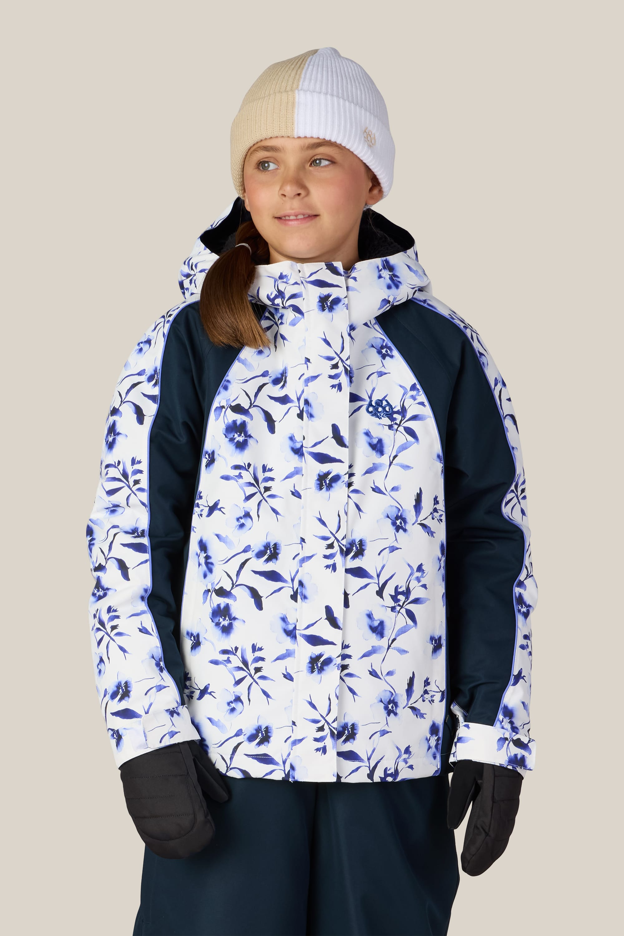 A young person wearing the 686 Girls’ Hologram™ Insulated Jacket, featuring a white and blue floral design from 686, paired with black gloves and a beige and white beanie, stands against a plain light background and looks slightly to the side.