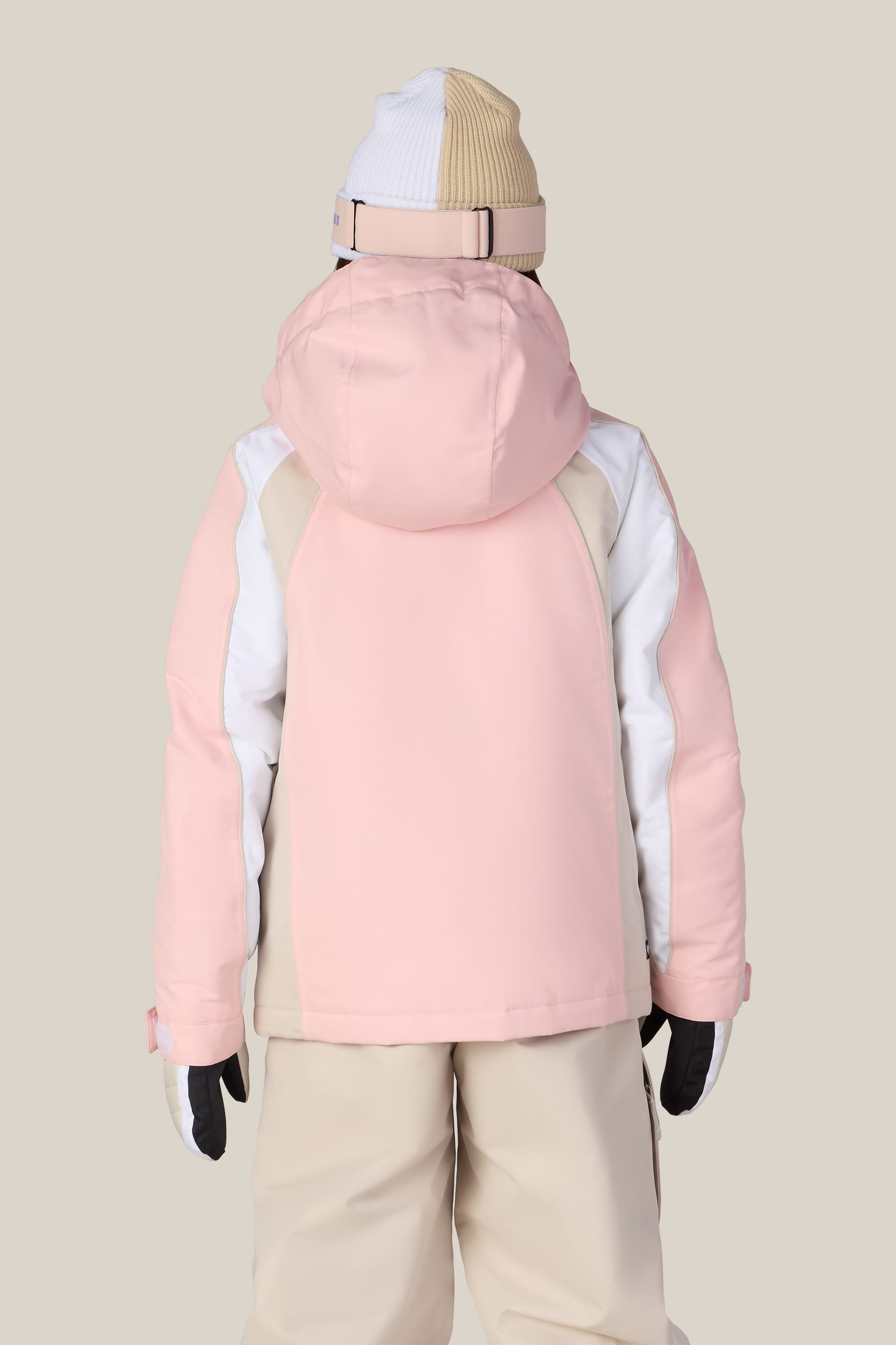 A person is facing away, wearing the 686 Girls’ Hologram™ Insulated Jacket in pink and white with a cozy polyester finish, beige snow pants, a white and beige knit hat, and black gloves, standing against a plain light background.