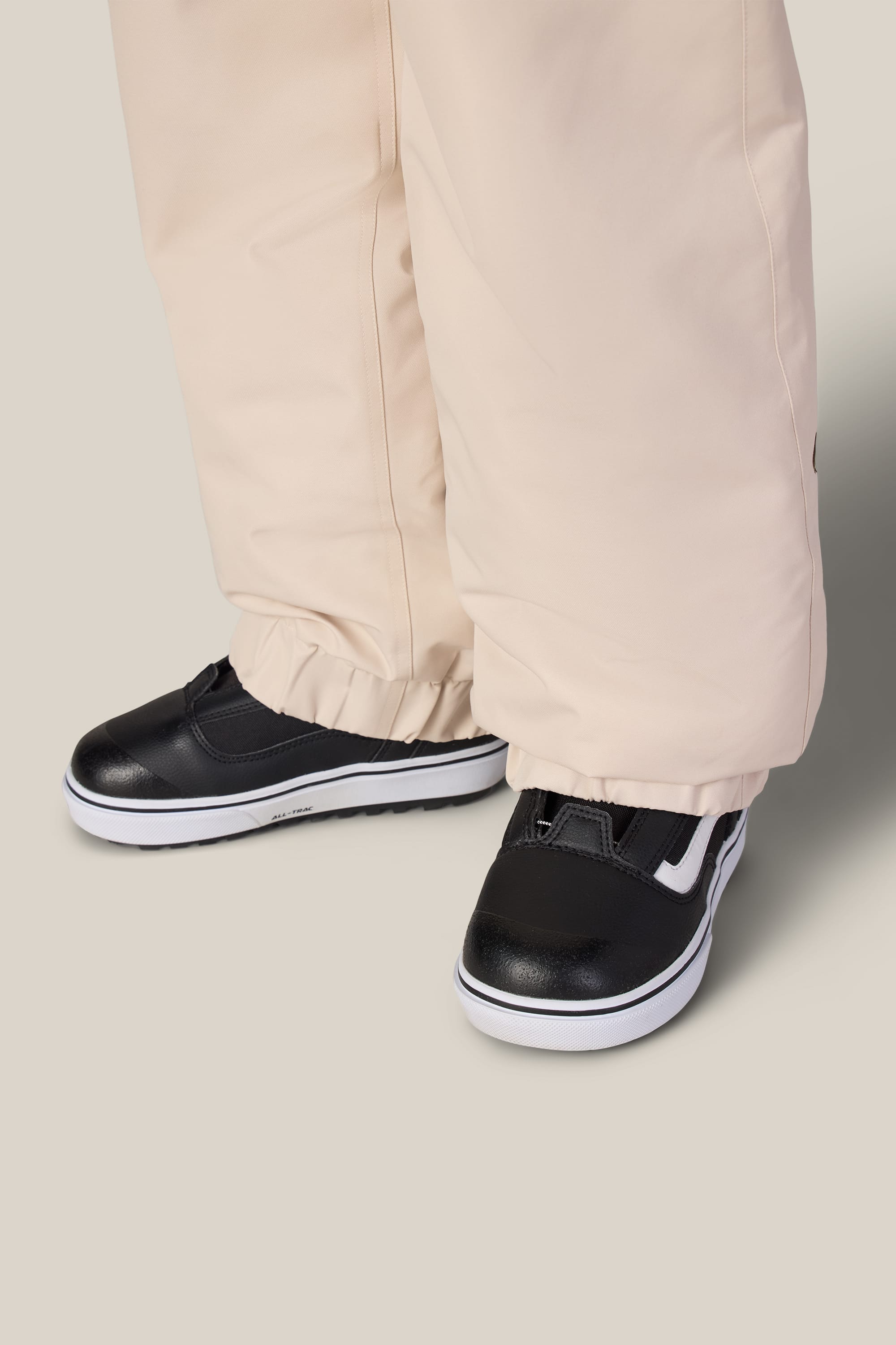 A person wearing the 686 Girls’ Outline™ Insulated Pant in beige from 686, featuring elastic cuffs, paired with black sneakers with white soles, stands on a light gray surface.