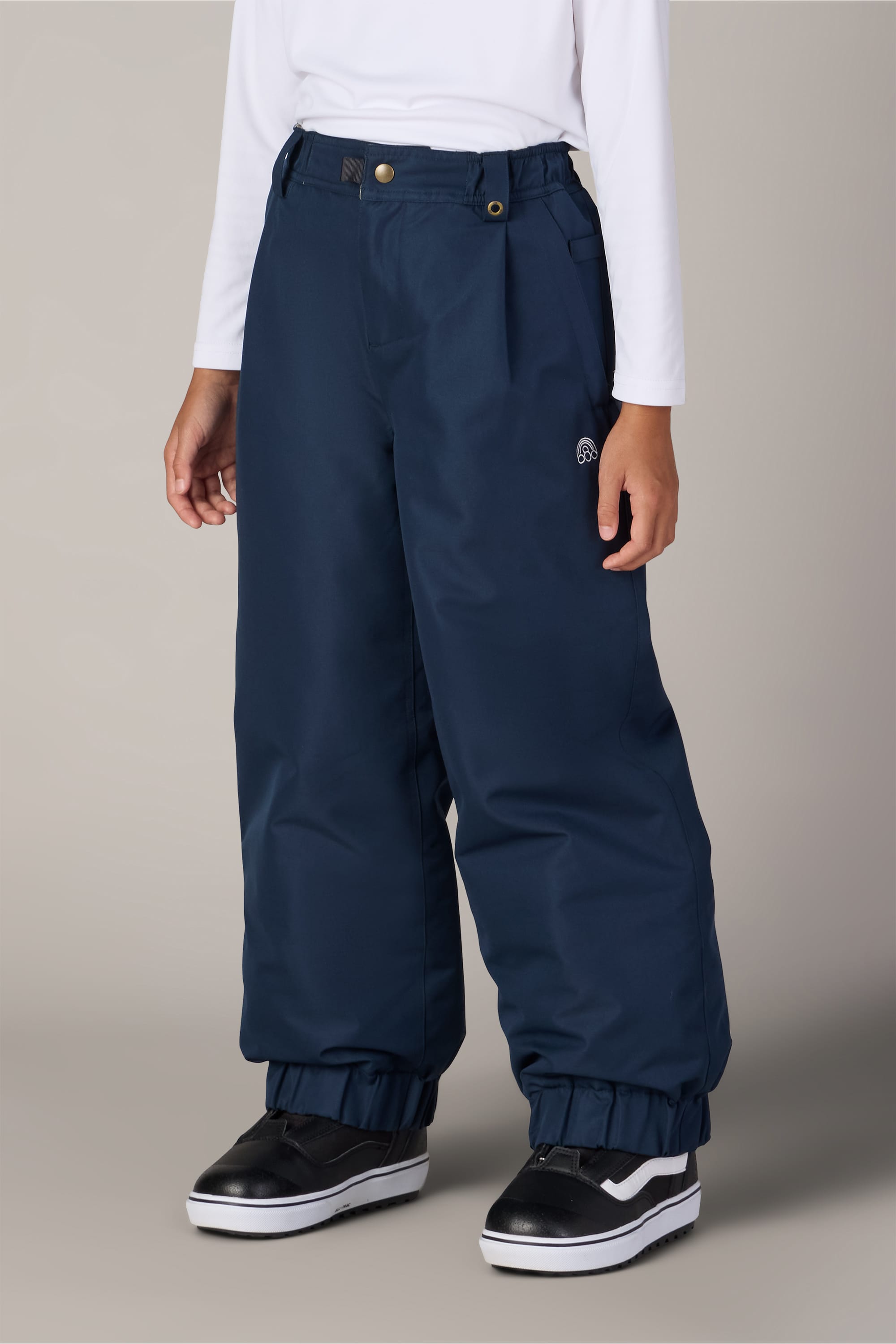 A child wearing a white long-sleeve shirt, the 686 Girls’ Outline™ Insulated Pant in navy blue by 686, and black and white sneakers stands against a plain light gray background.