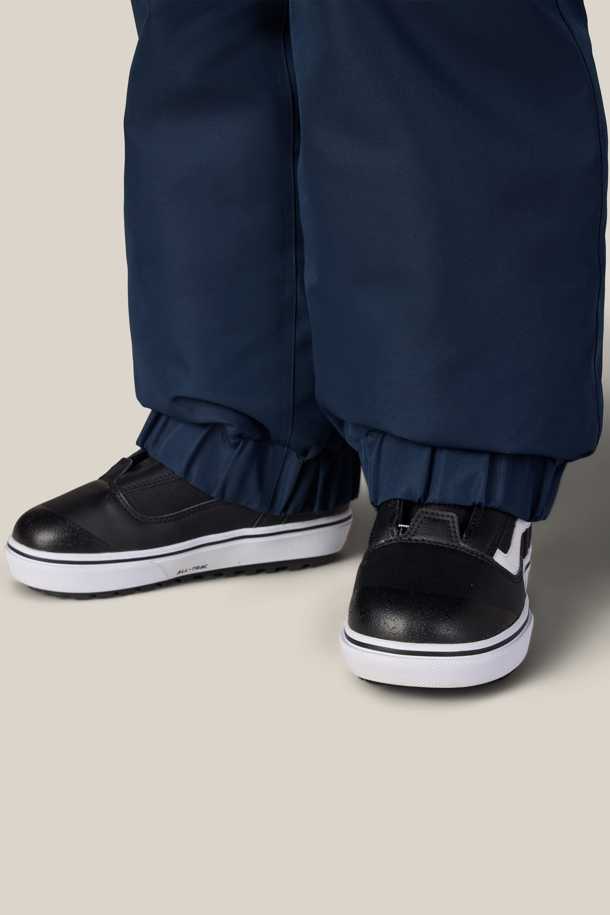 Close-up of a person wearing black and white sneakers and the 686 Girls’ Outline™ Insulated Pant by 686, featuring elasticized cuffs over the shoes, standing on a light-colored surface.