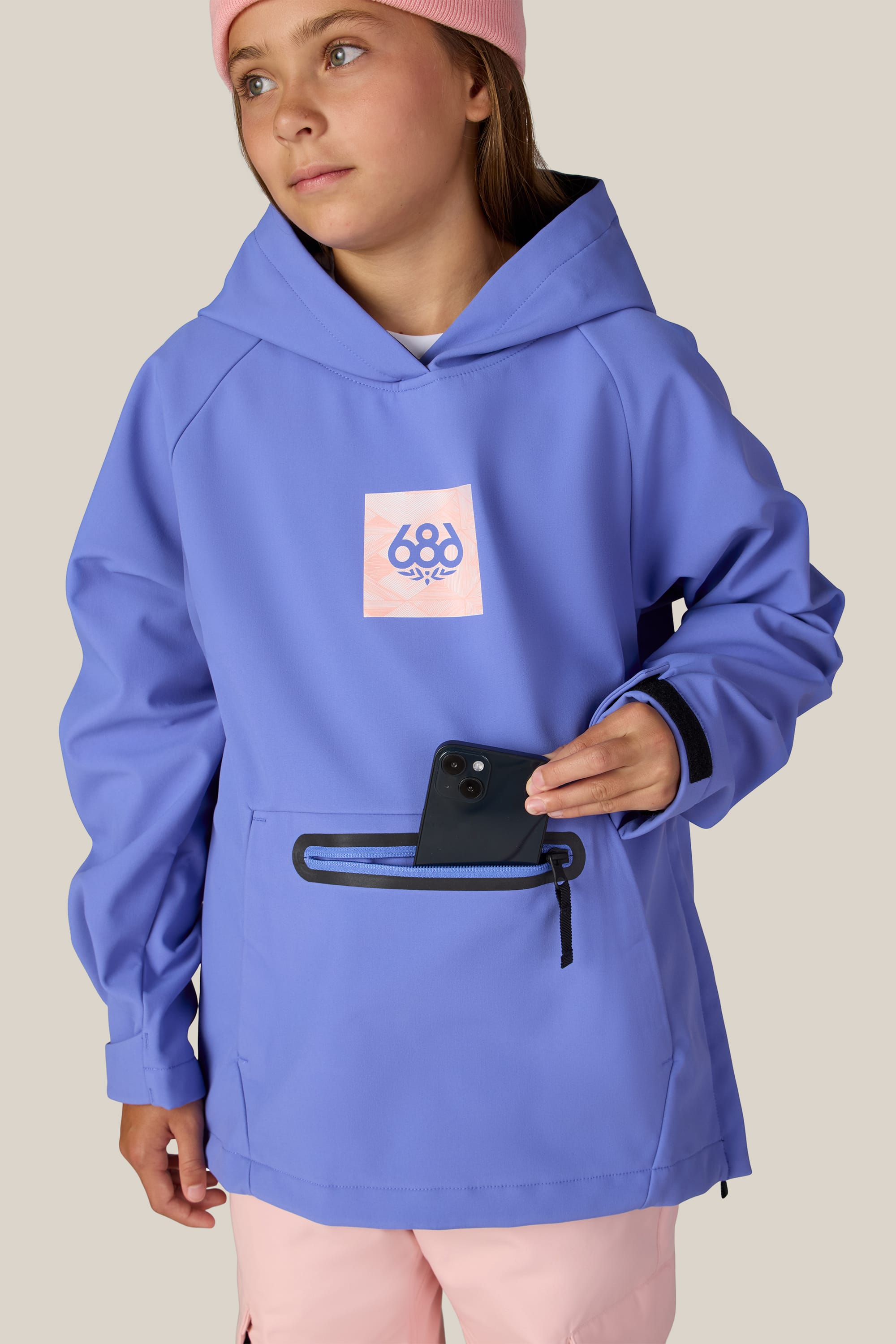 A child wearing the 686 Youth Waterproof Hoody in blue from 686, along with a pink beanie, puts a black smartphone into the front zippered pocket of their hoodie. The background is plain beige.