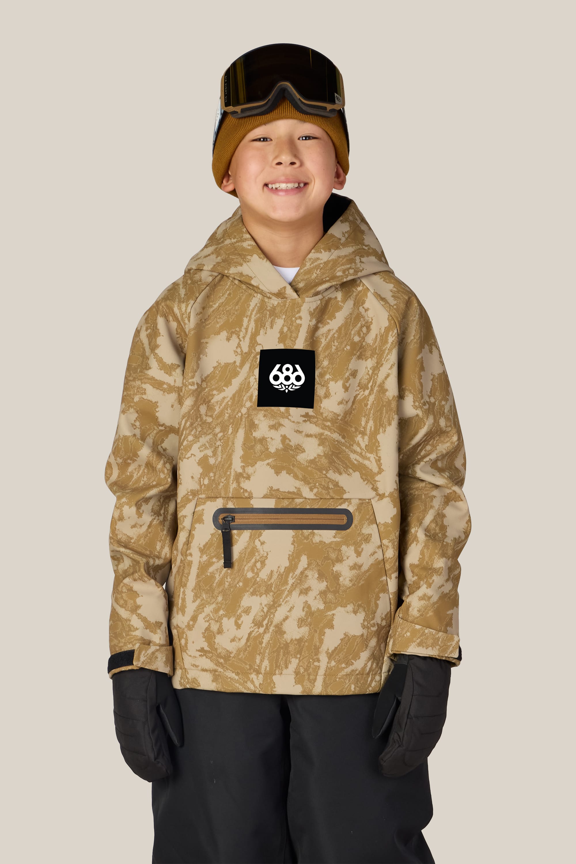 A smiling child stands against a plain beige background, dressed in a tan camo winter jacket layered over the 686 Youth Waterproof Hoody, paired with black snow pants, gloves, a brown beanie, and goggles perched on their head.