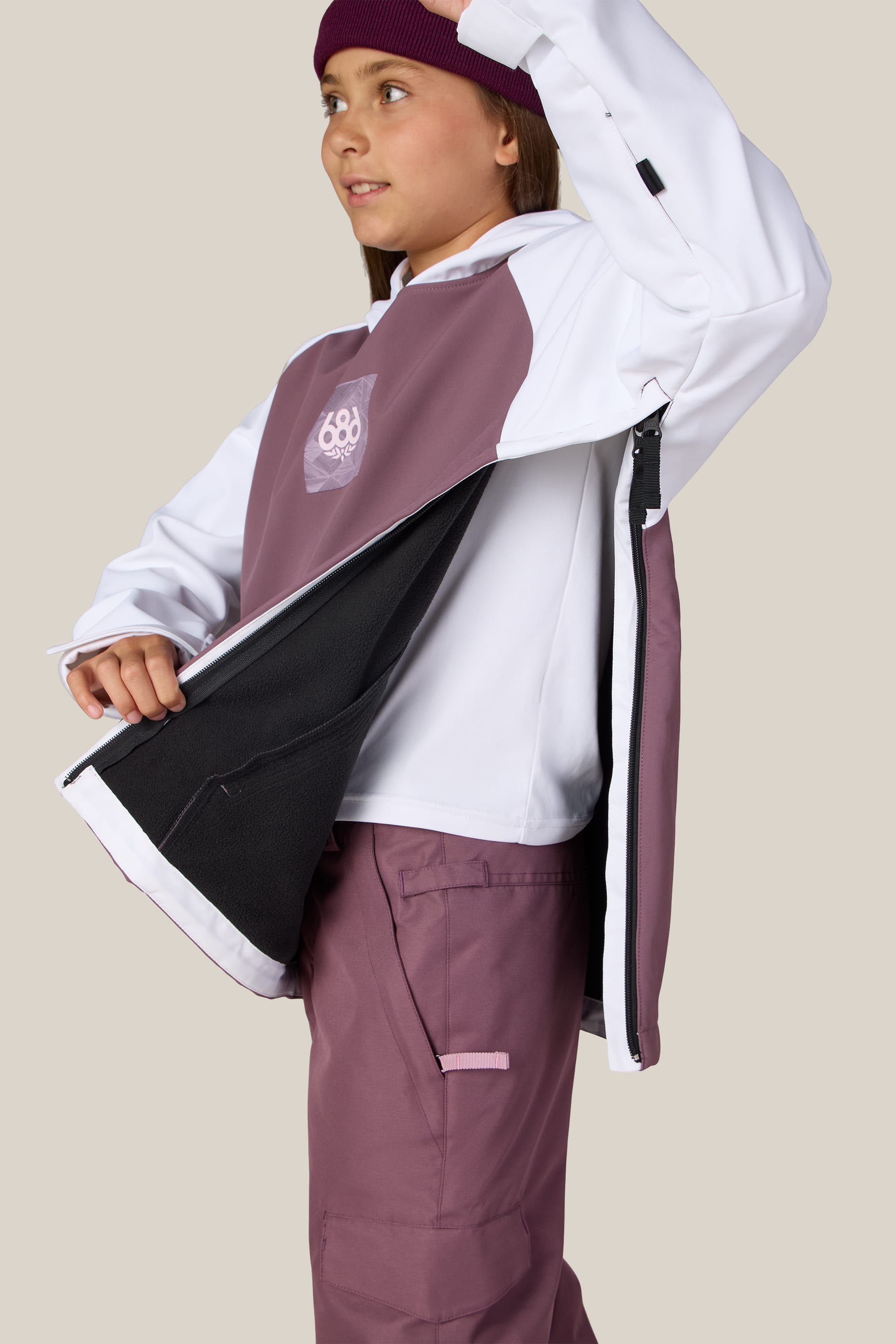 A person wearing the 686 Youth Waterproof Hoody in white and mauve polyester, paired with matching pants and a maroon beanie, is unzipping the side against a beige background.