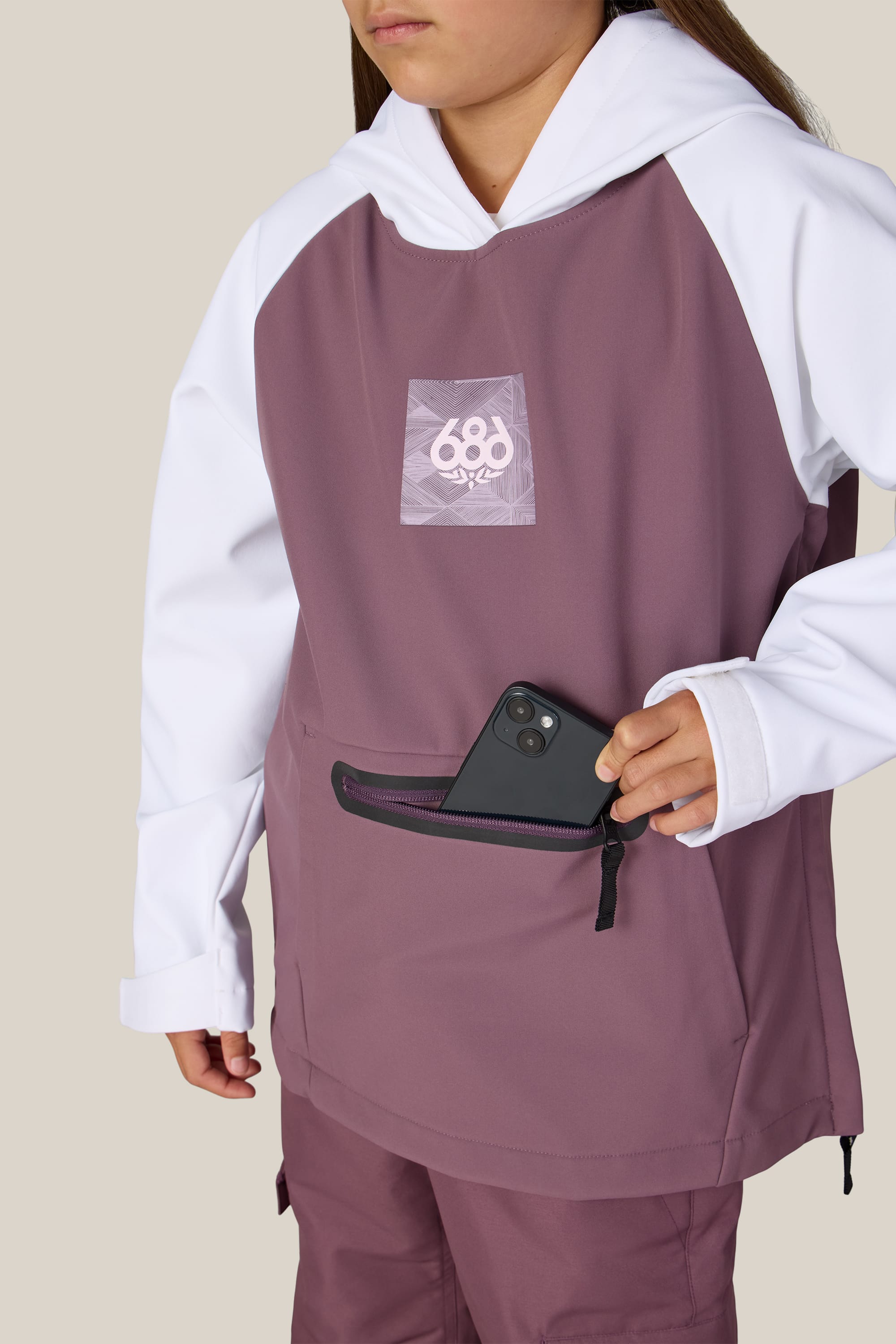 Wearing the 686 Youth Waterproof Hoody in maroon and white polyester, which features a white 686 logo on the chest, a person partially out of frame places a black smartphone into the front zippered pocket.