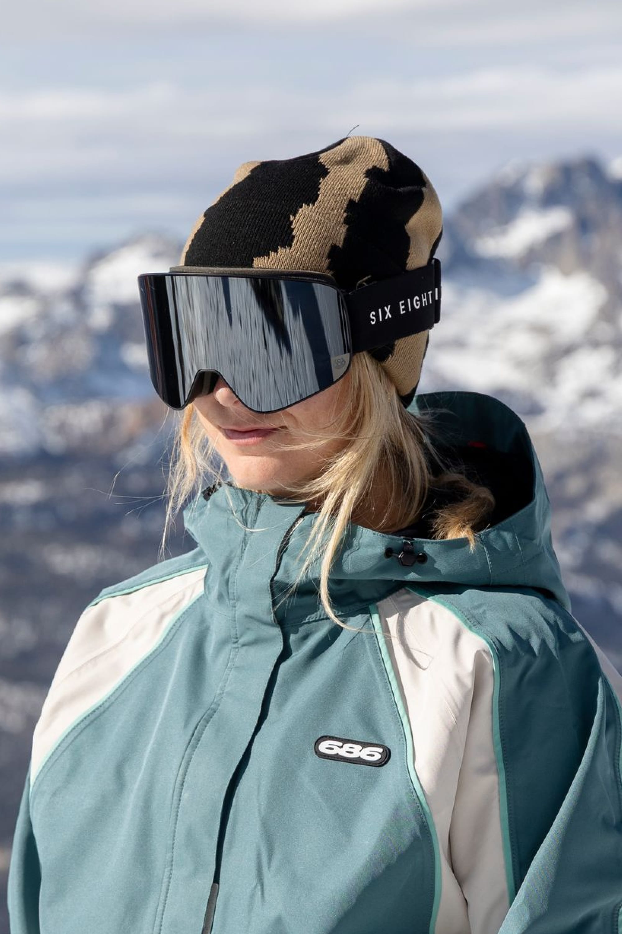 A person in a blue and white jacket, wearing the 686 Torn Beanie by 686 and large ski goggles, stands outdoors with snowy mountains blurred in the background.