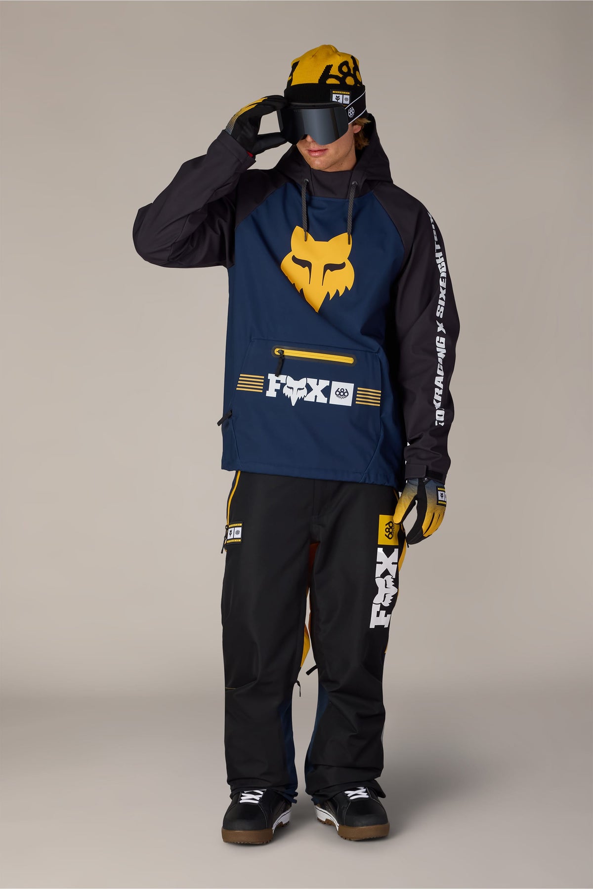 A person wearing a 686 Men’s Fox Racing Waterproof Hoody, matching pants, beanie, and goggles.