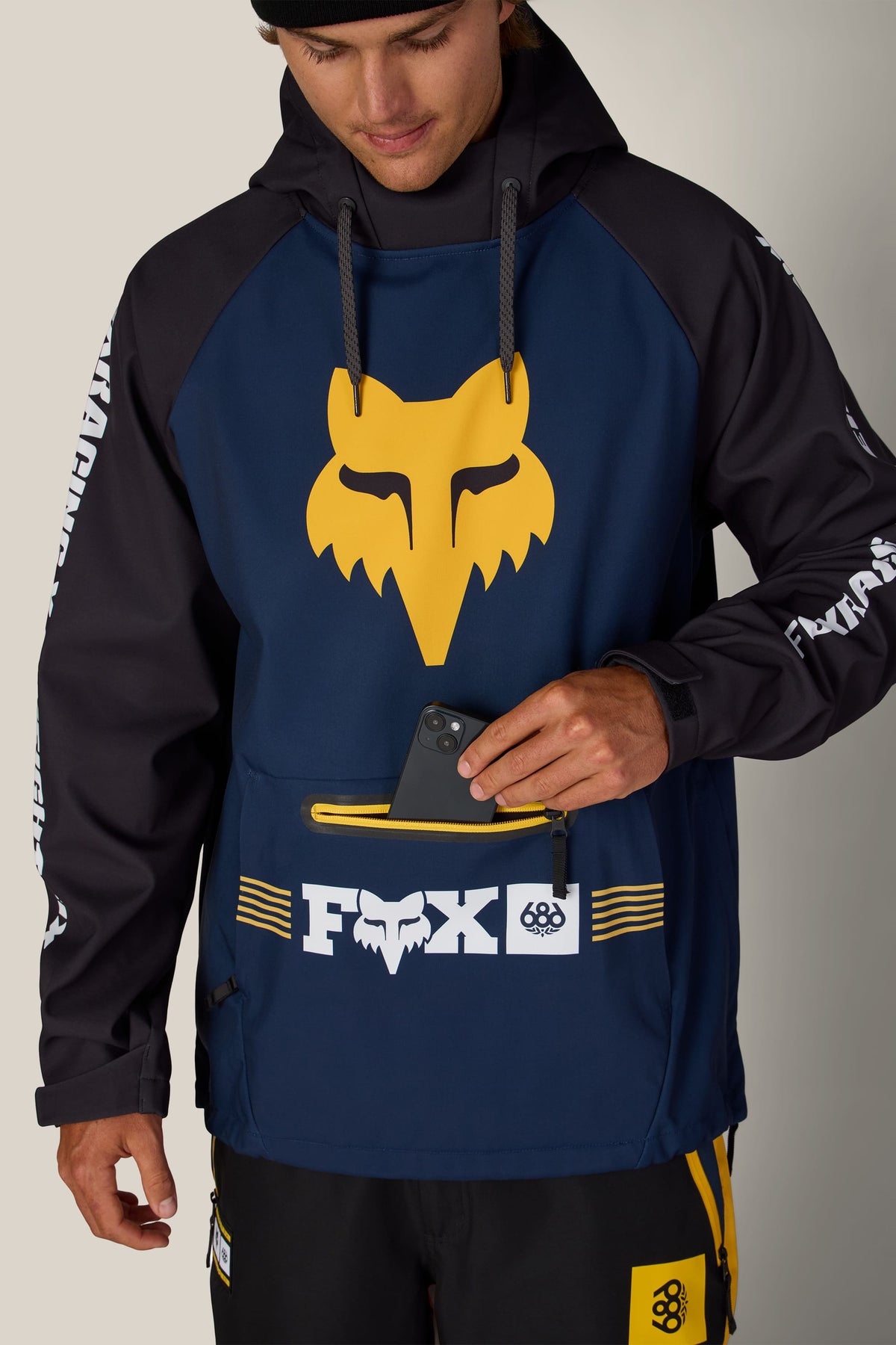 A person wearing a 686 Men’s Fox Racing Waterproof Hoody with a zippered front pocket holding a smartphone.