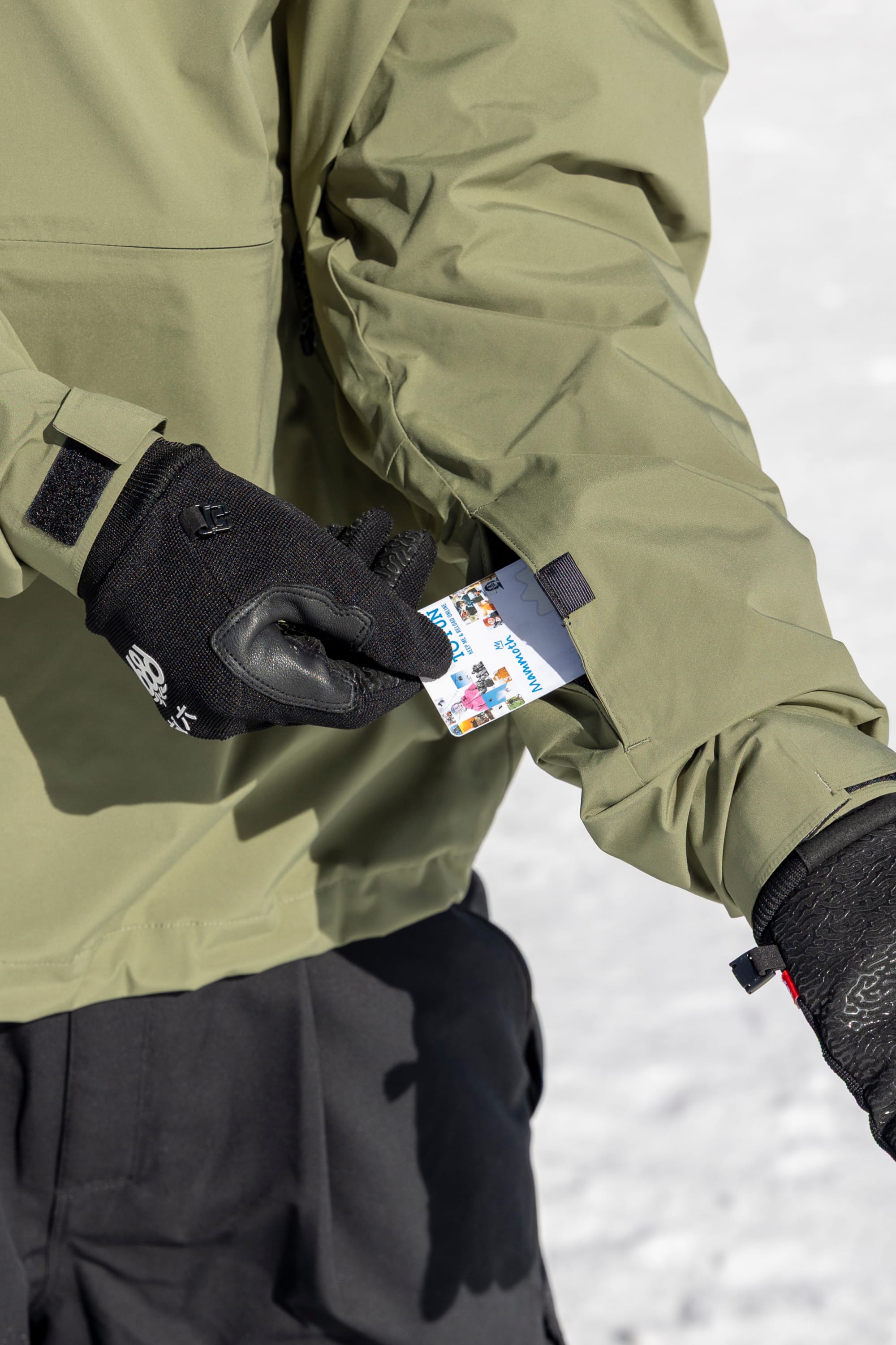 Wearing the 686 Men’s WINDSTOPPER® Pullover Hoody by GORE-TEX® LABS and black gloves, a person pulls a ski pass from their sleeve pocket against a snowy backdrop.