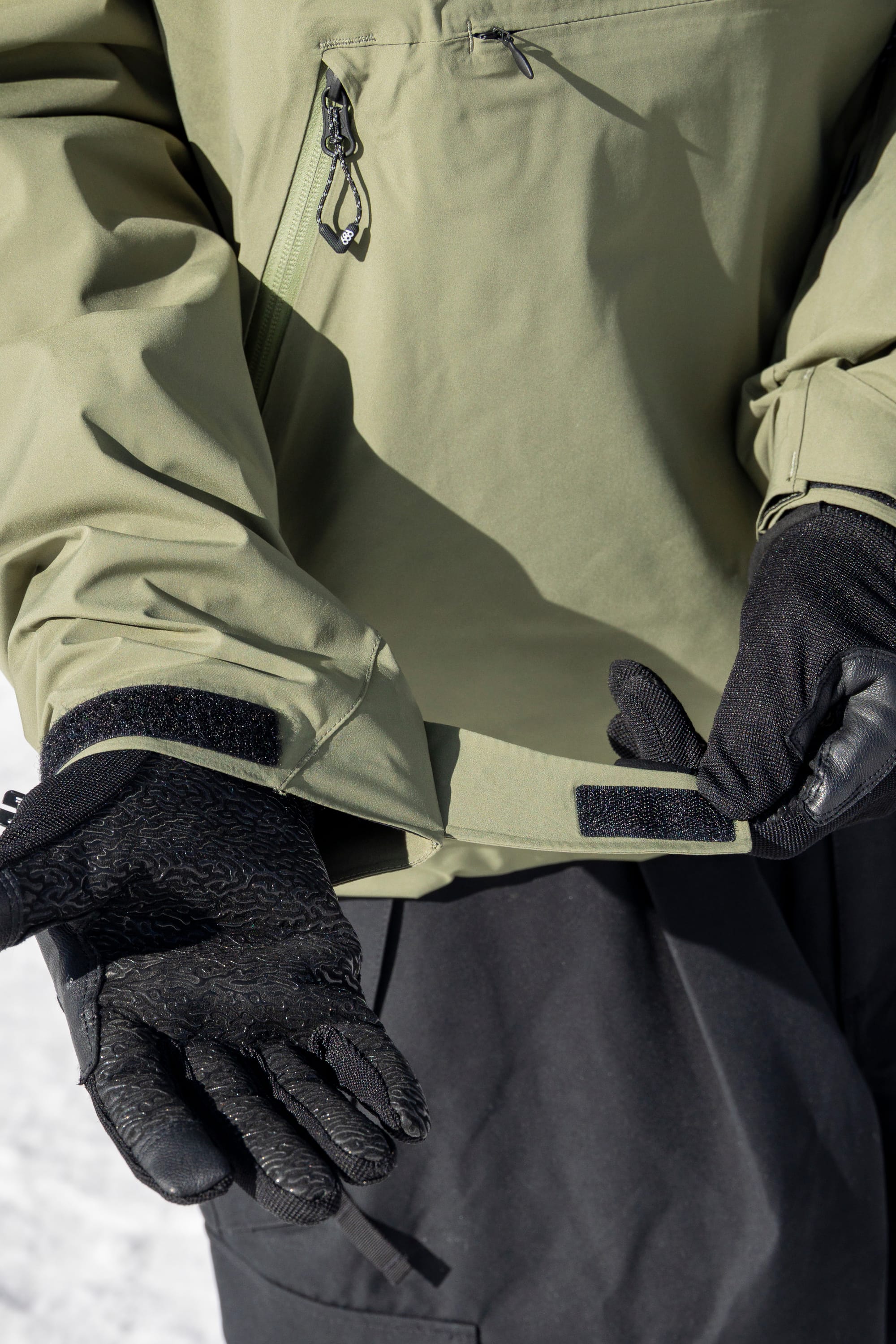 A person wearing black gloves and the 686 Men’s WINDSTOPPER® Pullover Hoody by GORE-TEX® LABS adjusts the Velcro strap on their jacket sleeve, with snow visible in the background.