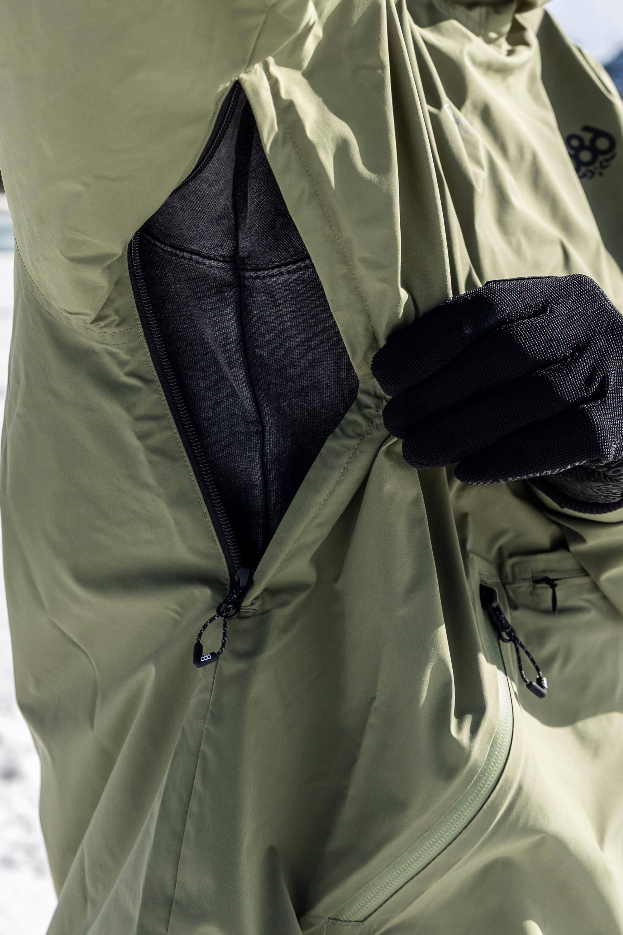 Wearing black gloves, a person unzips the armpit vent of the olive green 686 Men’s WINDSTOPPER® Pullover Hoody by GORE-TEX® LABS, revealing a dark inner layer, with snow visible in the background.