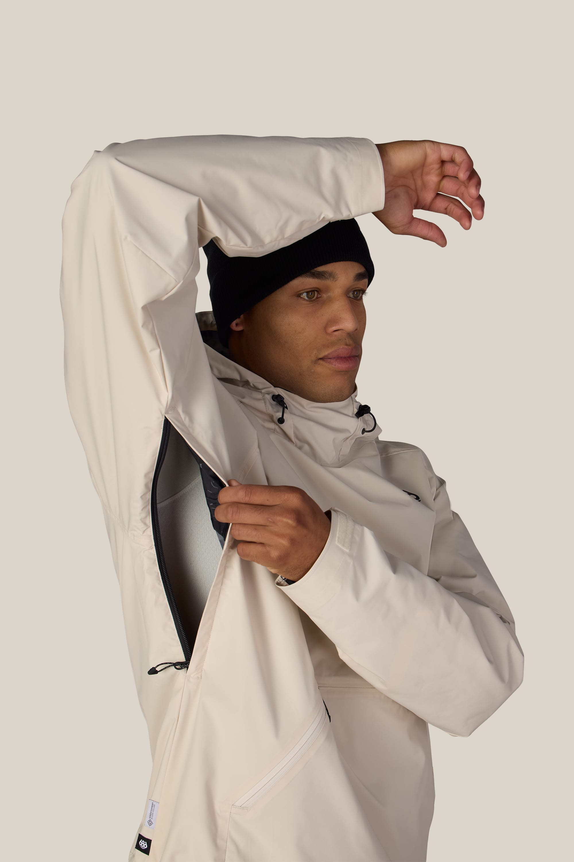 Wearing the 686 Men’s WINDSTOPPER® Pullover Hoody by GORE-TEX® LABS, a person in a cream jacket and black beanie lifts their arm to reveal the open underarm zipper for ventilation. This stylish men’s hoody by 686 blends modern design with breathability on a plain, light-colored background.