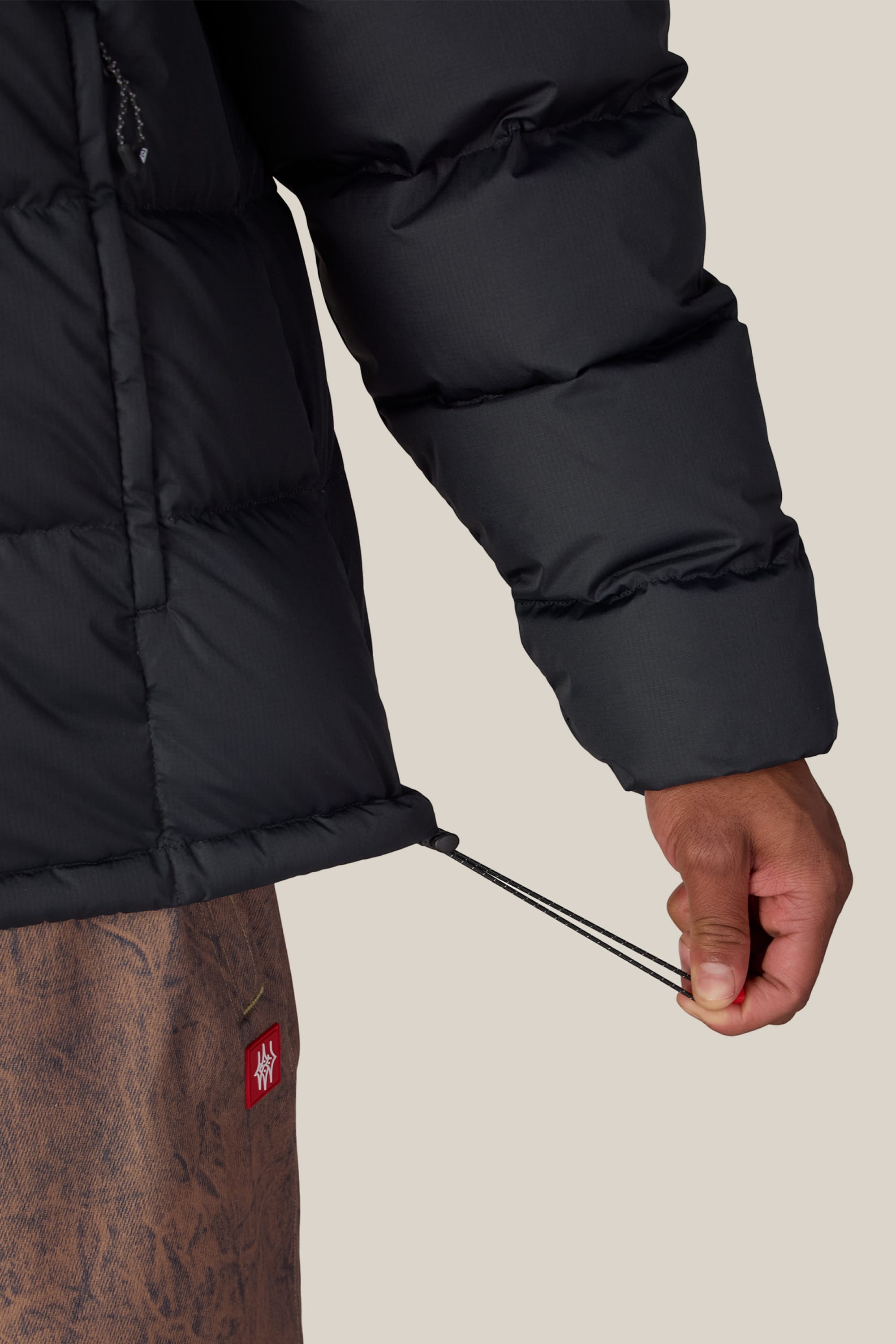 A person adjusts the drawstring at the hem of the 686 Men’s WINDSTOPPER® Pyro™ Down Puffer Jacket by GORE-TEX® LABS in black, paired with brown pants featuring a red and white tag, against a plain light background.