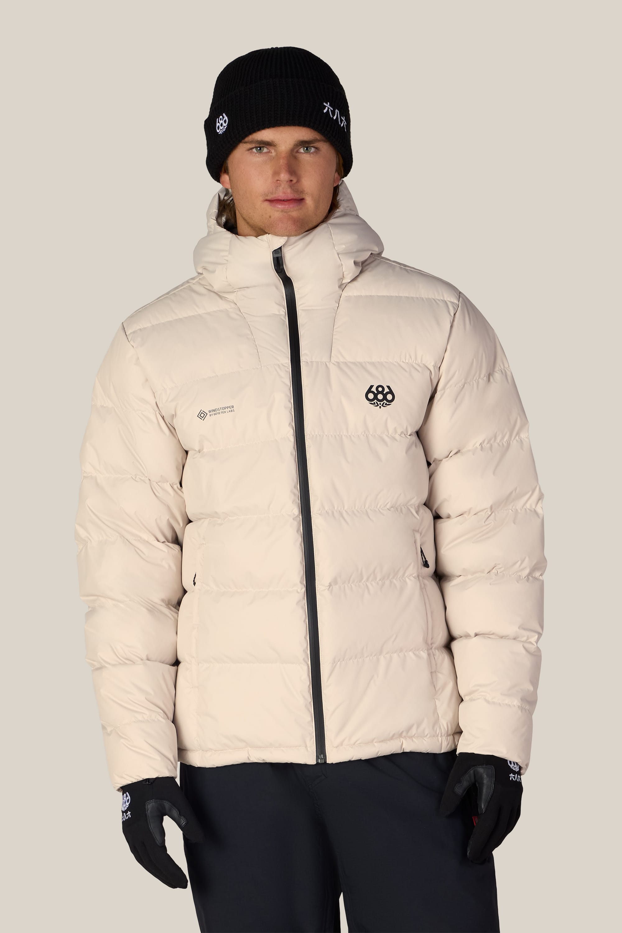 A person stands facing forward against a plain light background wearing the 686 Men’s WINDSTOPPER® Pyro™ Down Puffer Jacket by GORE-TEX® LABS in white, paired with black gloves and a black beanie.