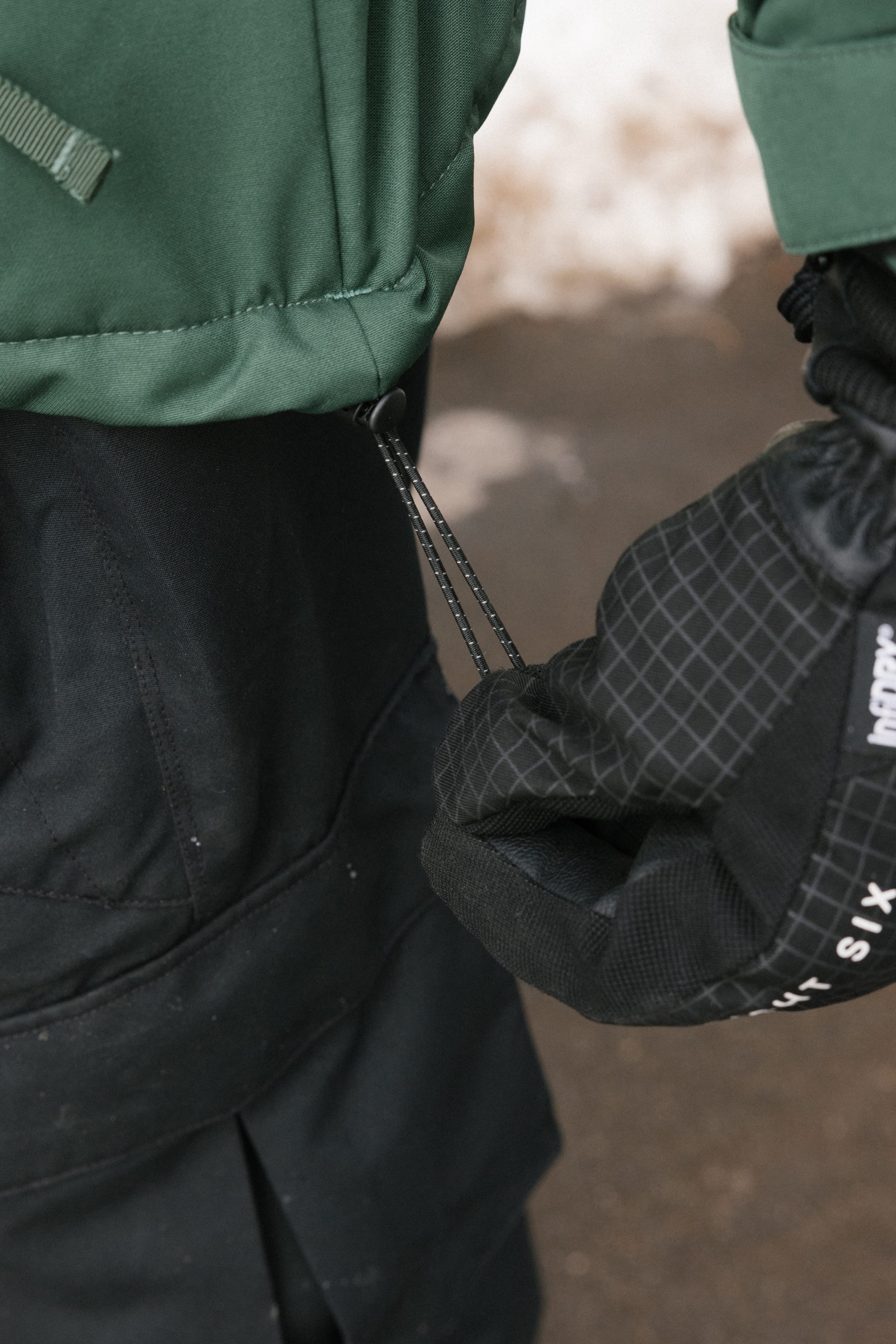 Wearing black gloves, a person pulls the drawstring on the 686 Men’s WINDSTOPPER® Guideline Insulated Jacket by GORE-TEX® LABS. The background is blurred, with hints of snow and pavement visible.