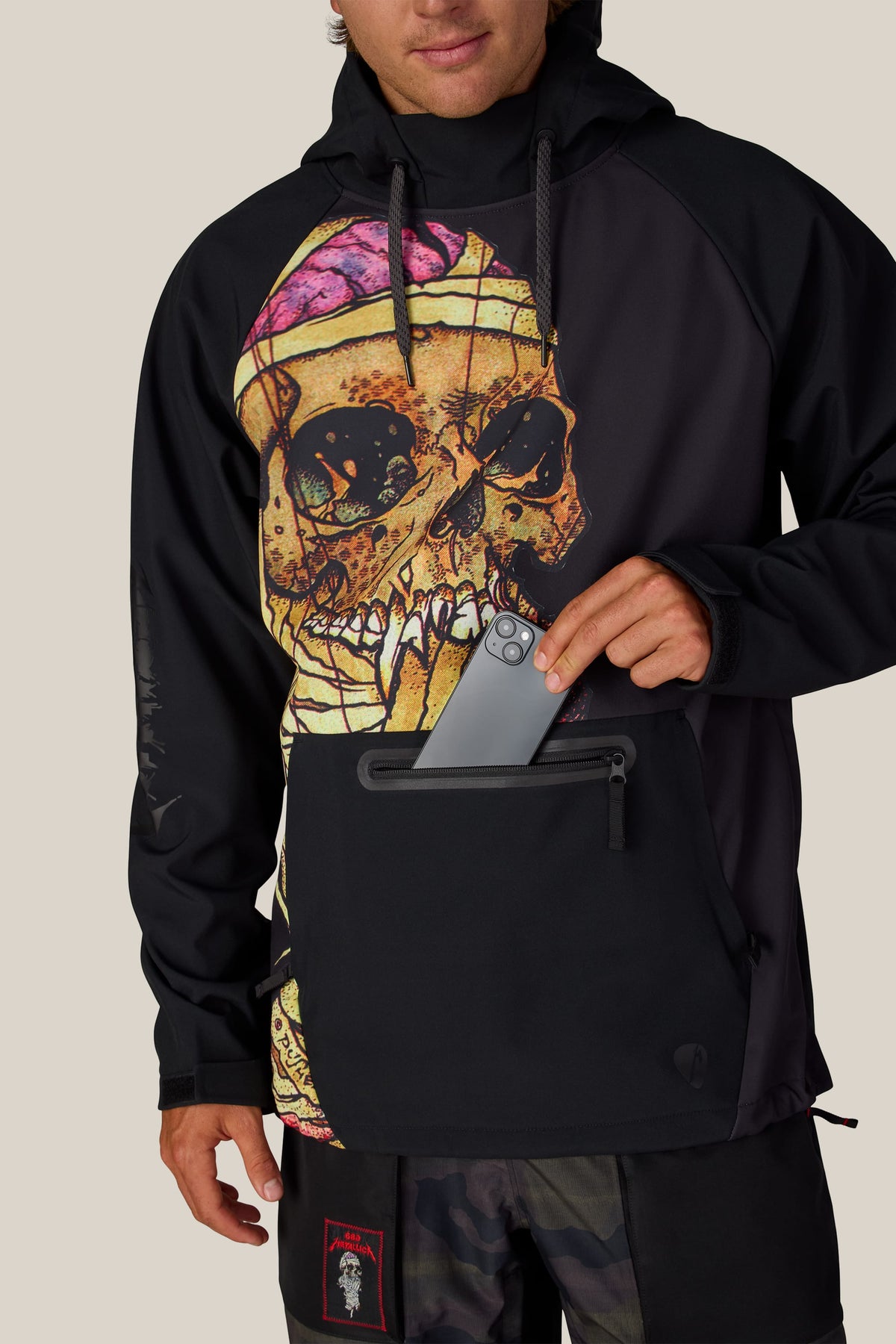 Person wearing a black 686 Men’s Metallica Waterproof Hoody featuring a large skull graphic on the chest and a zippered front pocket containing a smartphone.