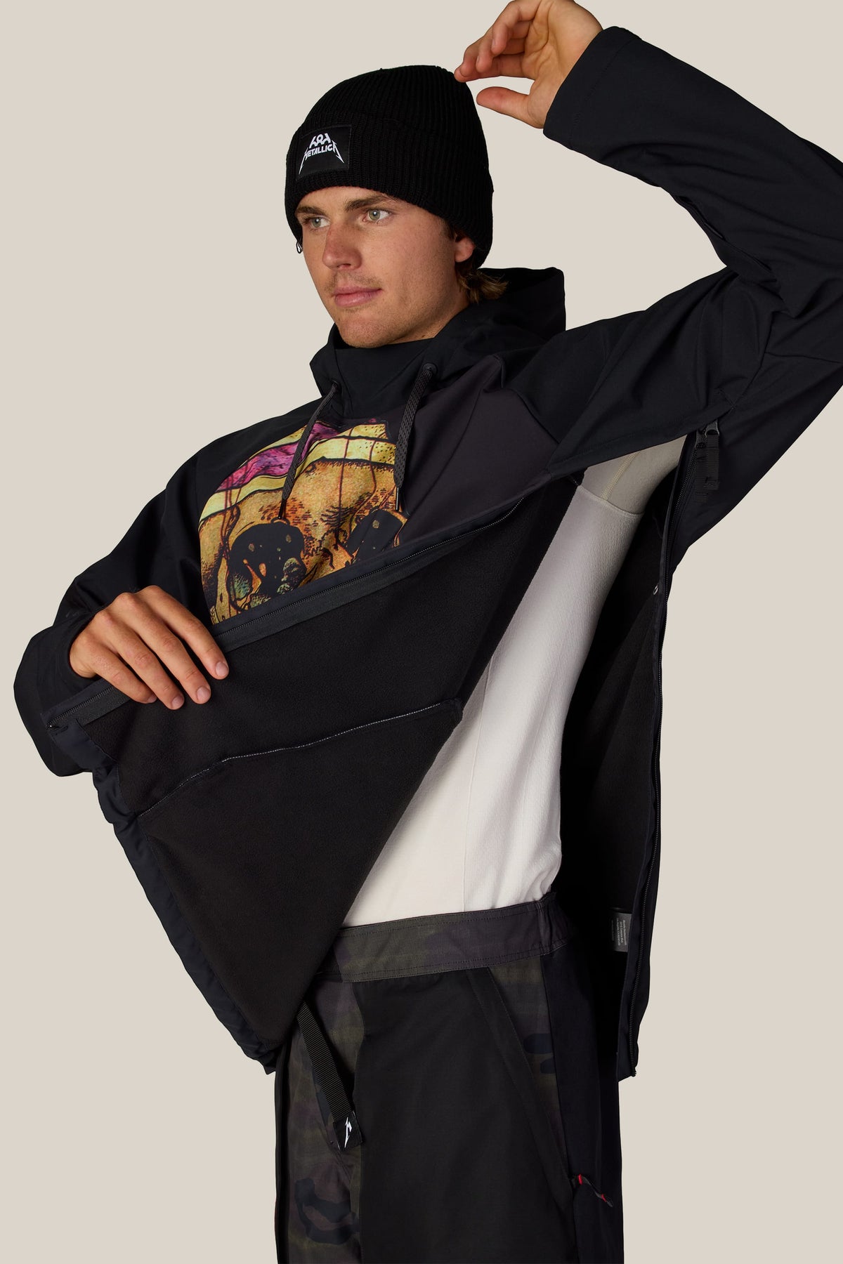 A person wearing a black beanie with a Metallica logo and a 686 Men’s Metallica Waterproof Hoody, partially unzipped to show the interior lining.