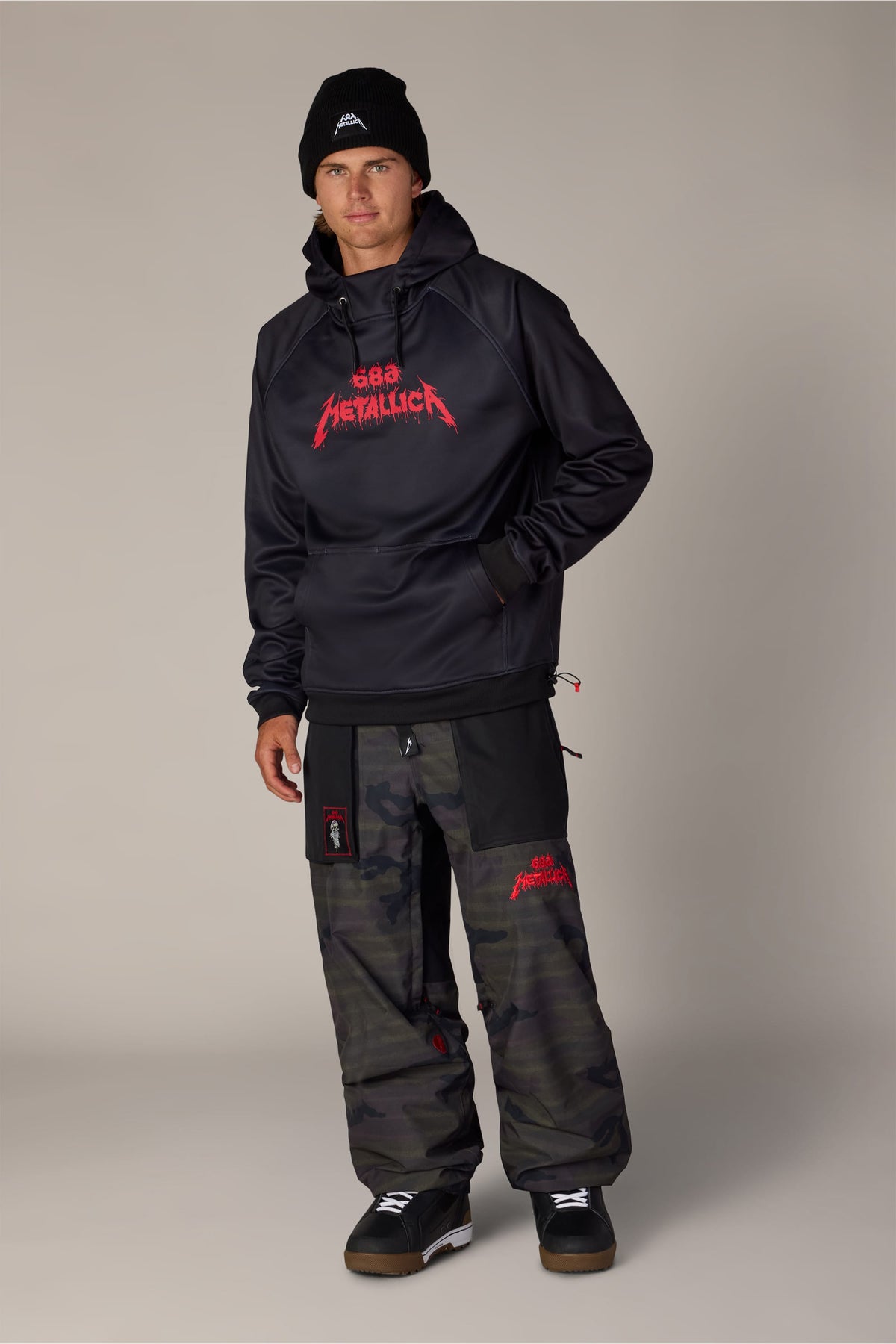 A person wearing a 686 Men’s Metallica Bonded Fleece Pullover Hoody, a black beanie, and dark camouflage snow pants.