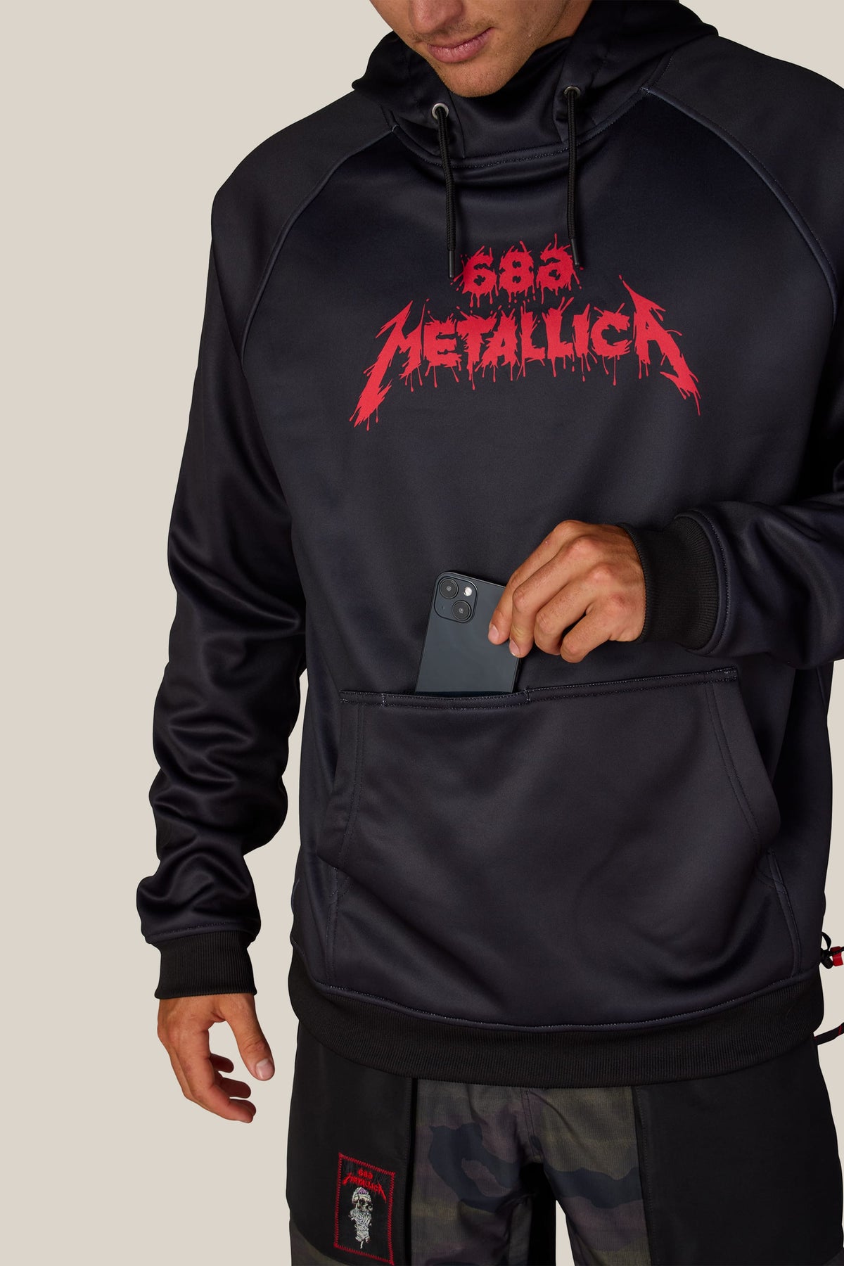 686 Men’s Metallica Bonded Fleece Pullover Hoody in black polyester, featuring a red Metallica logo on the chest and a hand sliding a smartphone into a dedicated front pocket.