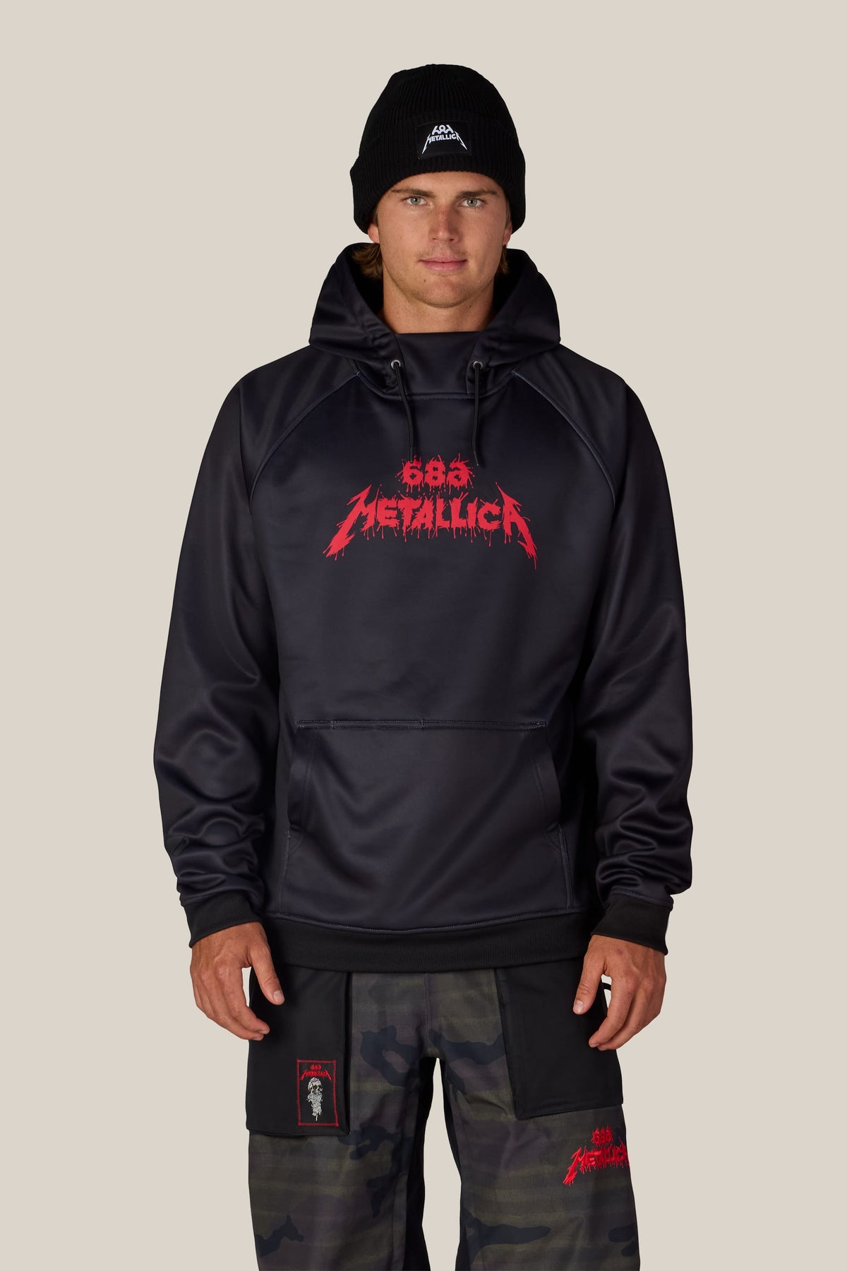 A person wears a black 686 Men’s Metallica Bonded Fleece Pullover Hoody with a red Metallica logo across the chest, a black beanie with a small logo, and camouflage pants.