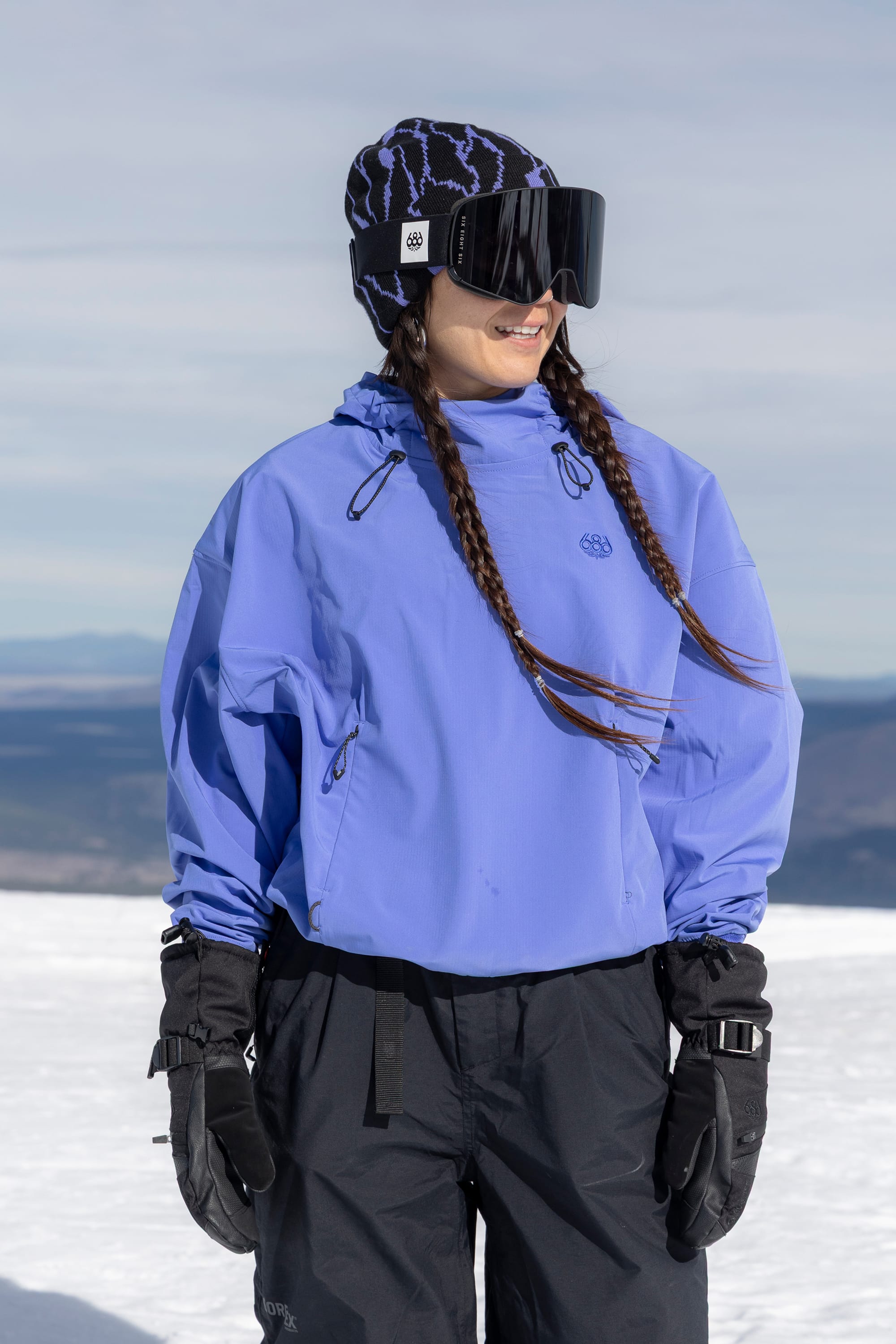 A woman wearing the 686 Women’s Waterproof Team Crop Hoody in blue, paired with black snow pants, gloves, large ski goggles, and a patterned beanie stands on the snow with braided hair, looking to the side. Mountains and a cloudy sky fill the background.