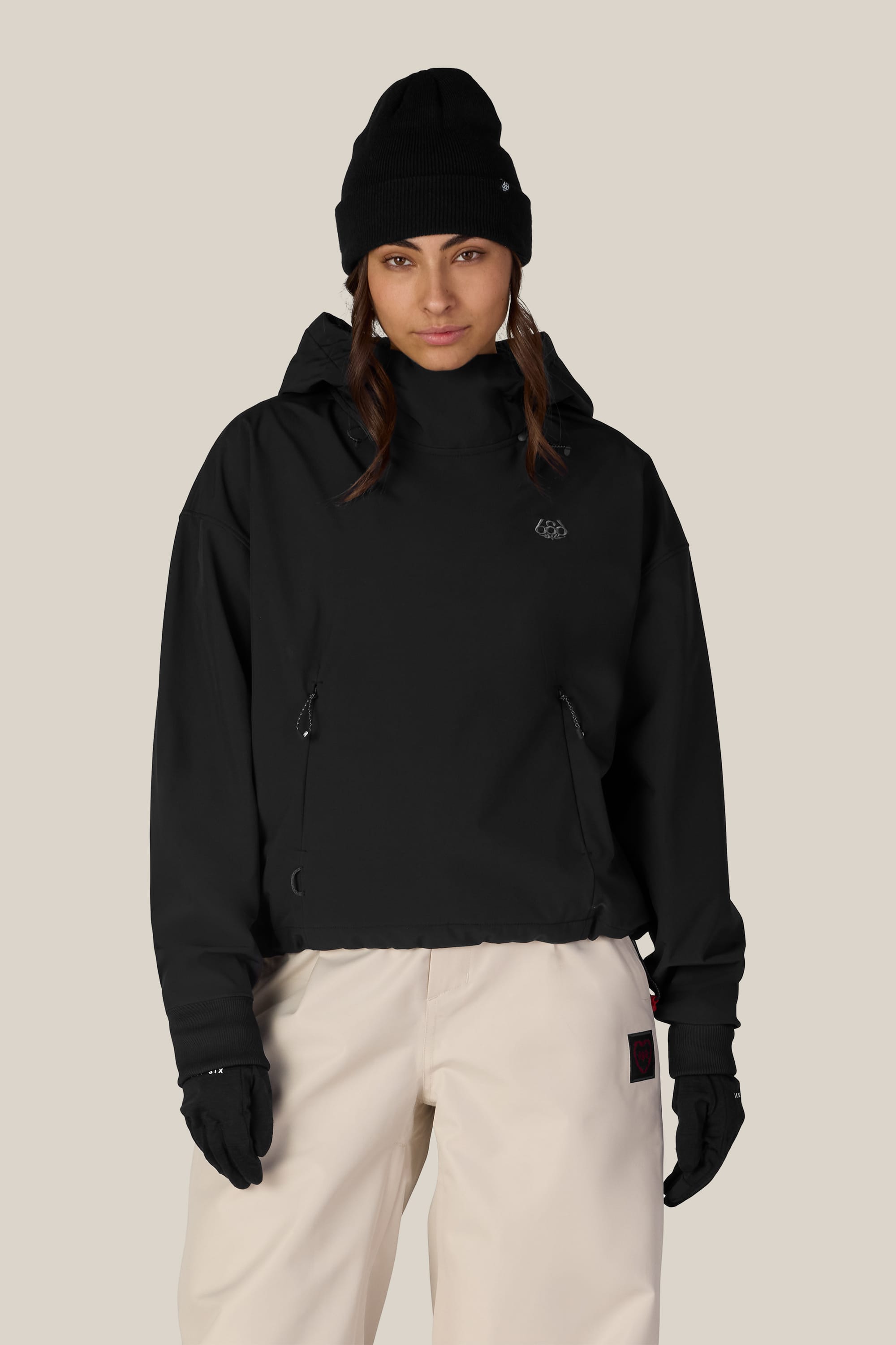 A person wearing a black beanie, black gloves, white pants, and the 686 Women’s Waterproof Team Crop Hoody stands facing forward against a plain beige background.