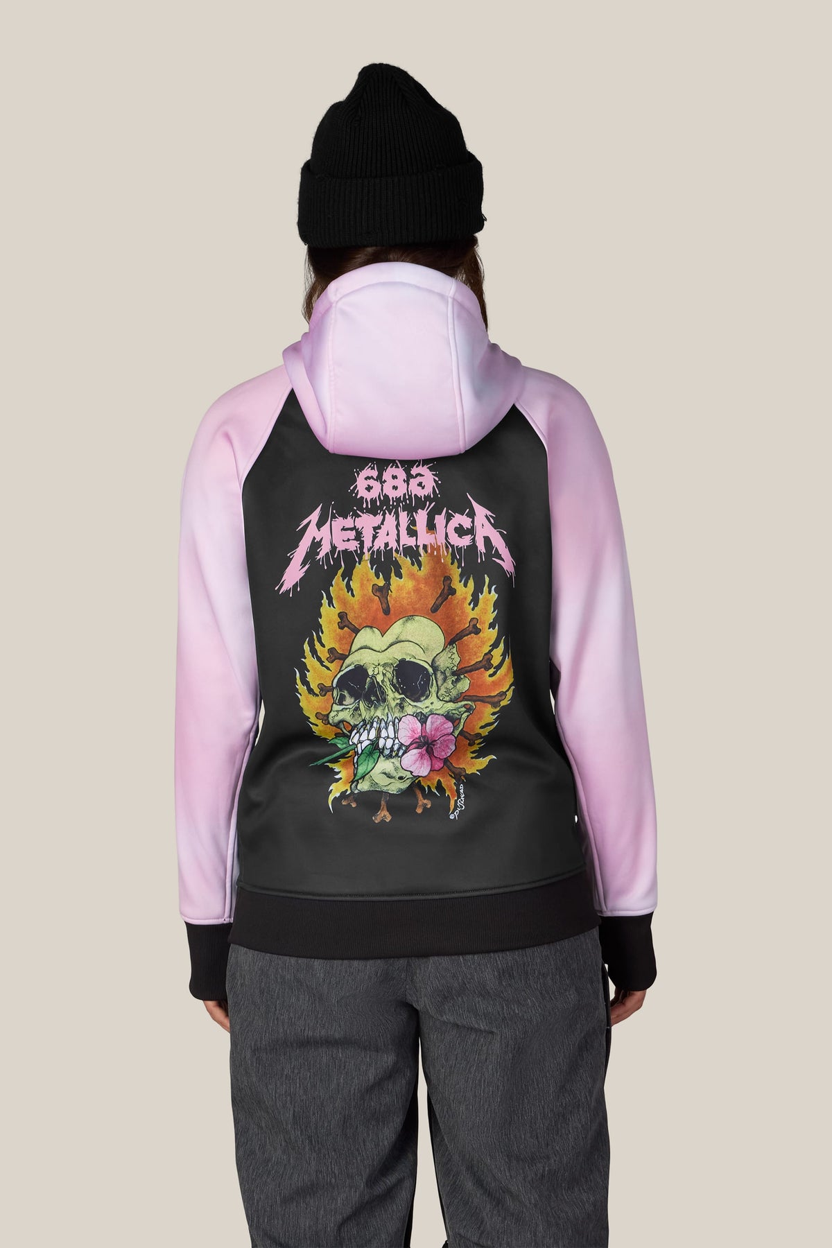 A person wearing a 686 Women’s Metallica Bonded Fleece Pullover Hoody featuring a black back panel with a Metallica graphic of a skull, flames, and a pink flower.
