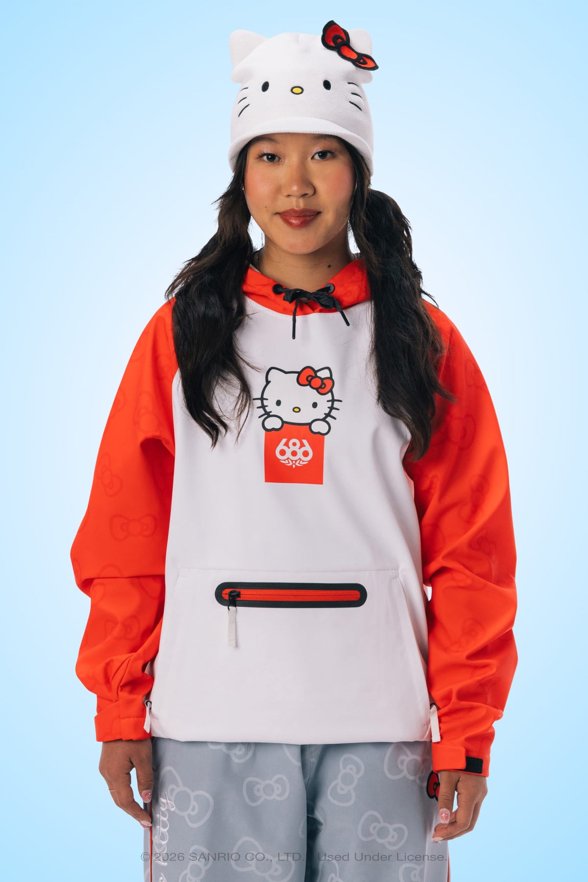 A person is wearing a white Hello Kitty hat, the 686 Women’s Hello Kitty® Waterproof Hoody by 686—a white and red woven hoodie with a zipper pocket—and light blue pants with subtle Hello Kitty patterns, standing against a light blue background.