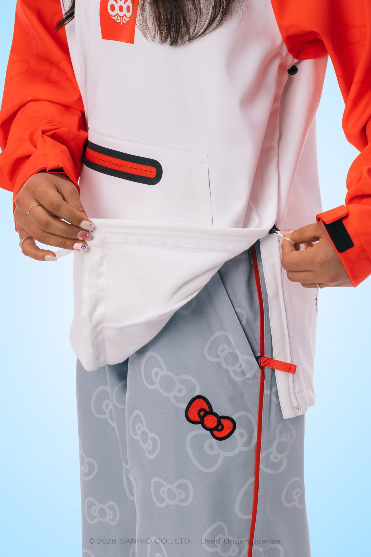 A person, visible only from the torso down, unzips the side of the 686 Women’s Hello Kitty® Waterproof Hoody—a white woven hoody with red sleeves—revealing gray pants decorated with white Hello Kitty bows and a single red Hello Kitty bow near the knee.
