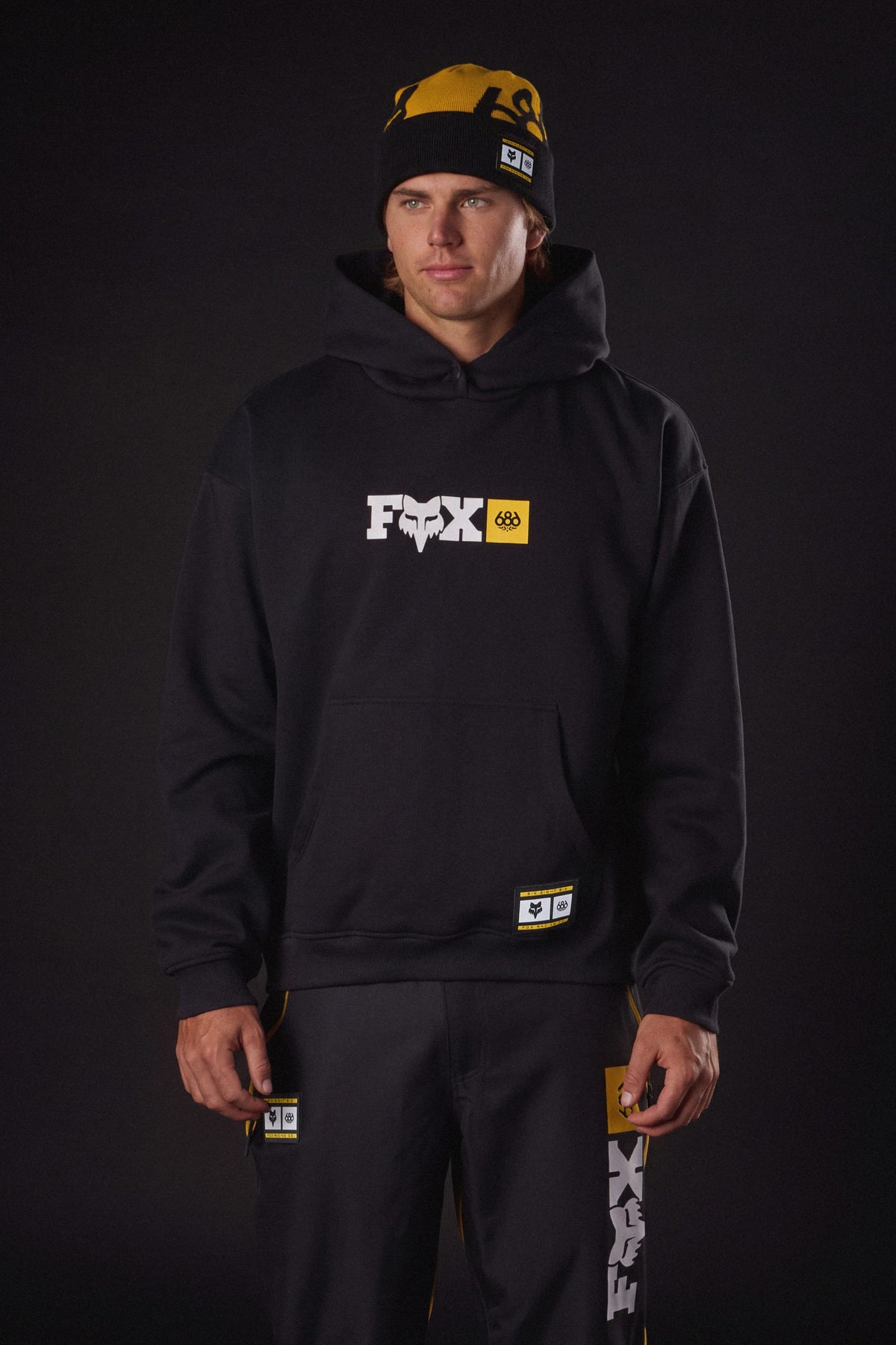 A man wearing a black 686 Men’s Fox Racing Premium Heavyweight Pullover Hoody, matching sweatpants, and a yellow and black beanie.