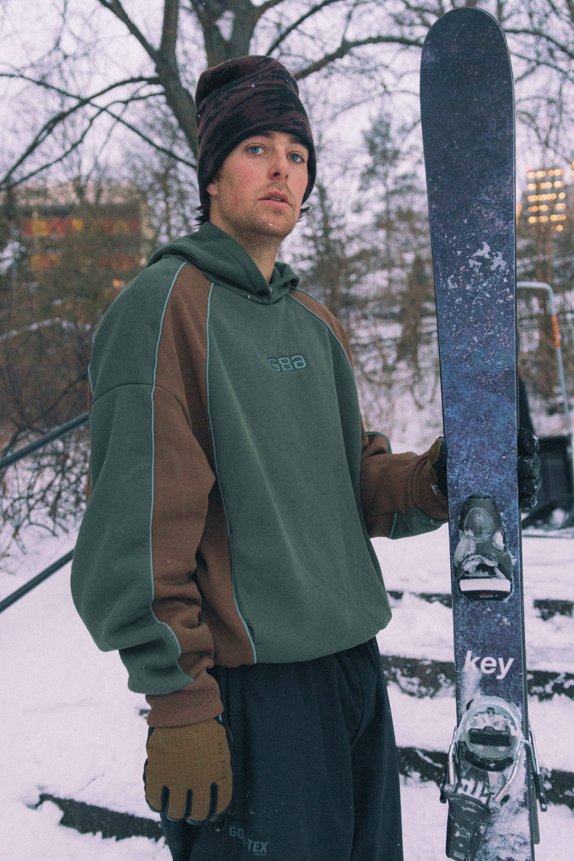 Wearing the 686 Legacy Premium Heavyweight Hoody, a man stands outdoors in the snow with gloves on, holding a pair of skis upright. Snow-covered trees and steps create a wintry backdrop.