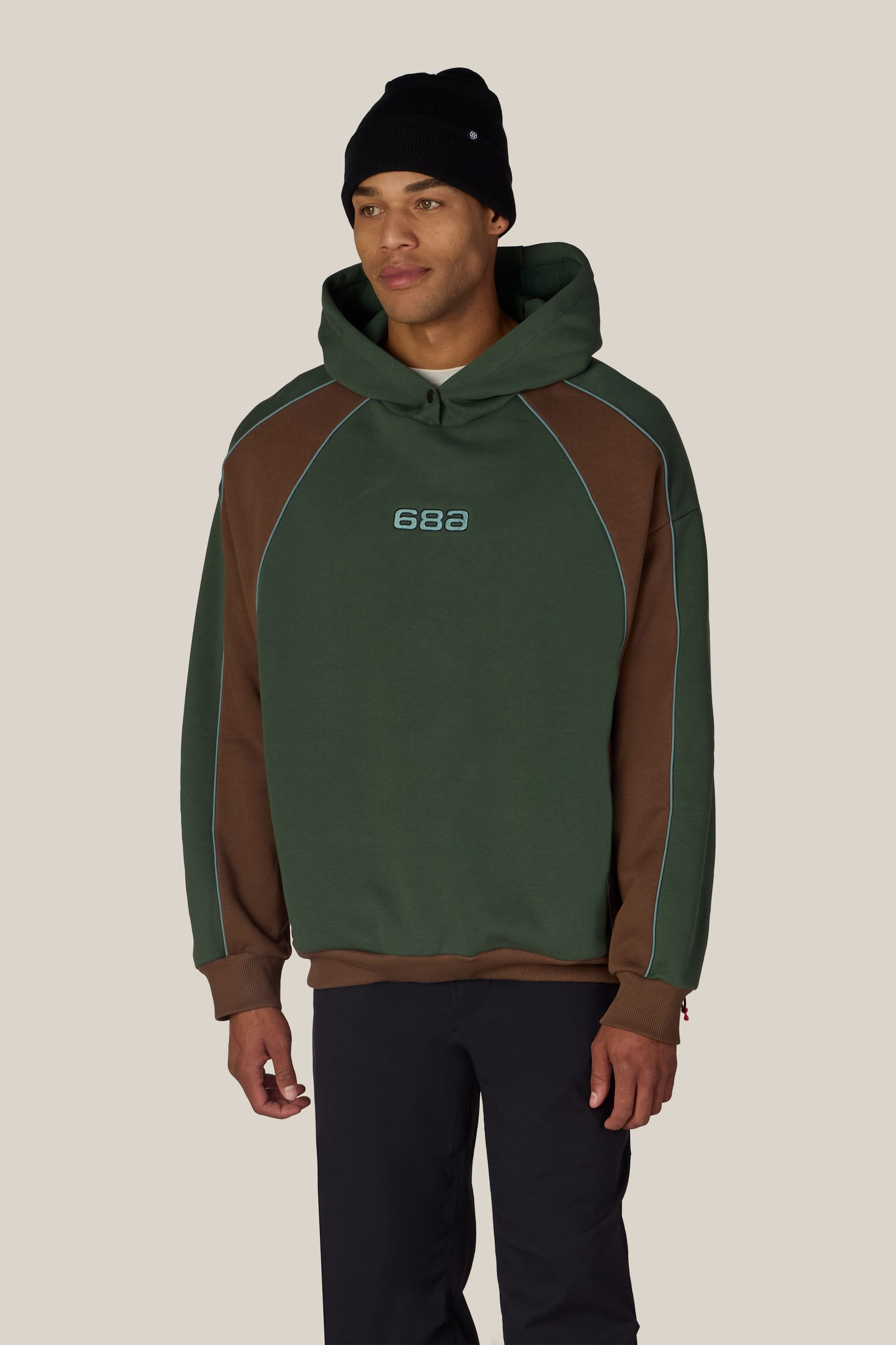 A man wearing the 686 Legacy Premium Heavyweight Hoody in green and brown, black pants, and a black beanie stands against a plain light background.