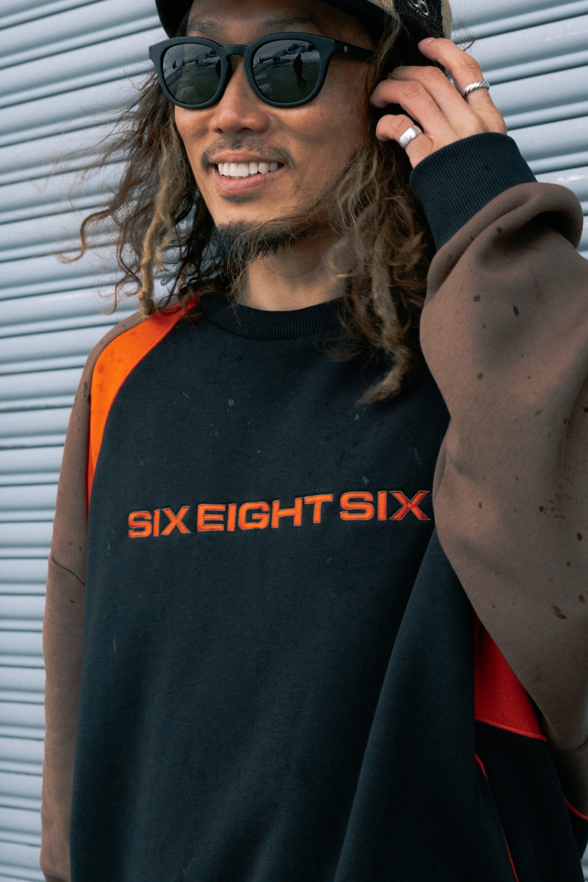 A man with long hair and sunglasses smiles as he stands in front of a corrugated metal wall, wearing the 686 Reserved® Premium Heavyweight Crewneck—a black and brown men’s sweatshirt featuring bold orange “SIX EIGHT SIX” lettering from 686.