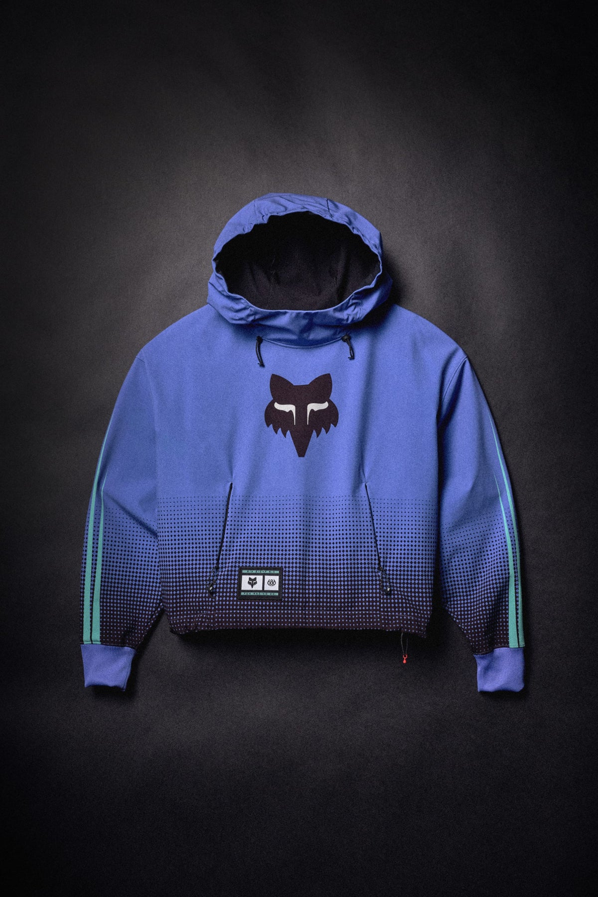 686 Women’s Fox Racing Waterproof Crop Hoody in a blue and black gradient, featuring a Fox head logo on the chest and a brand patch near the hem.