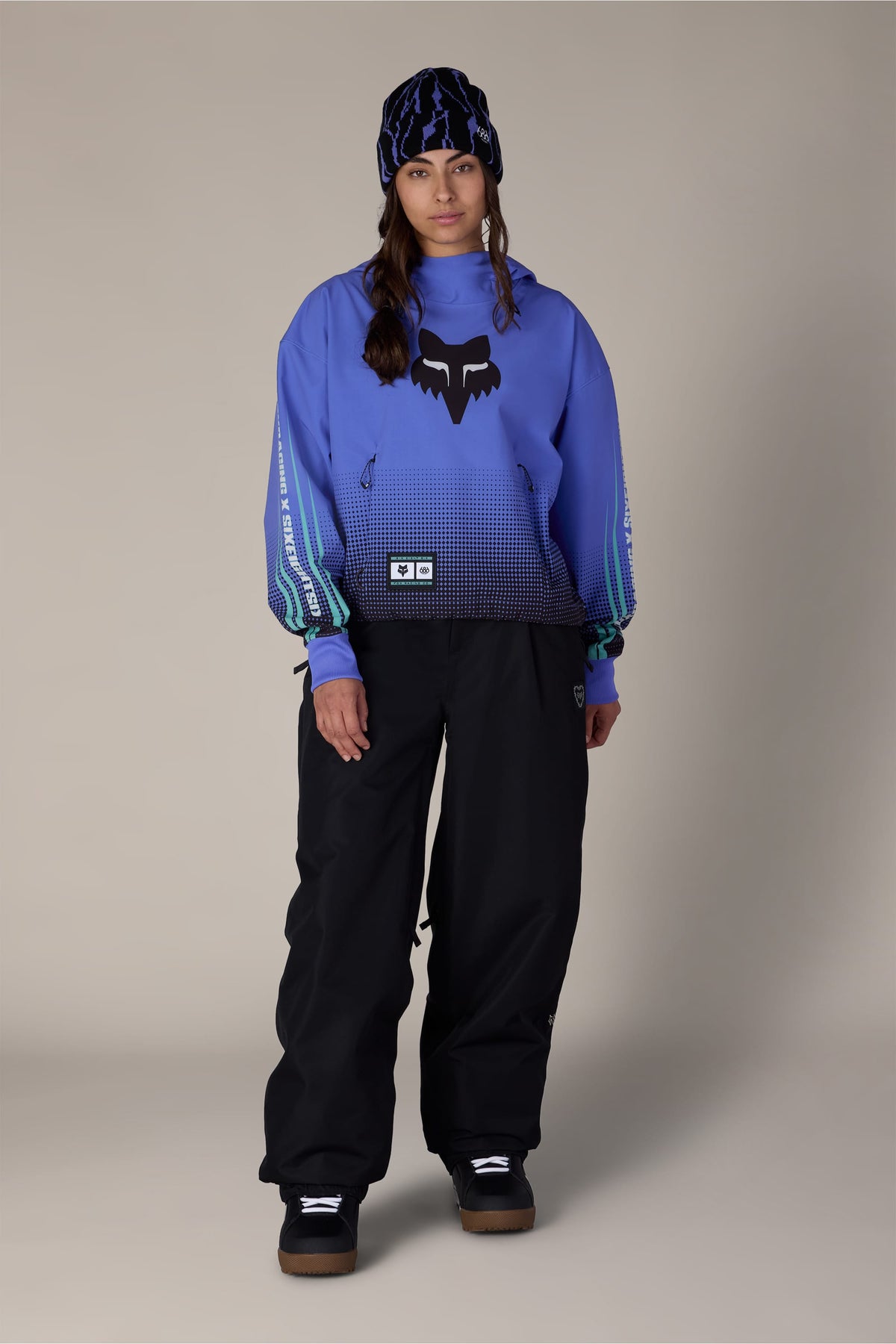 A person wearing a 686 Women’s Fox Racing Waterproof Crop Hoody, black snow pants, and a patterned beanie.