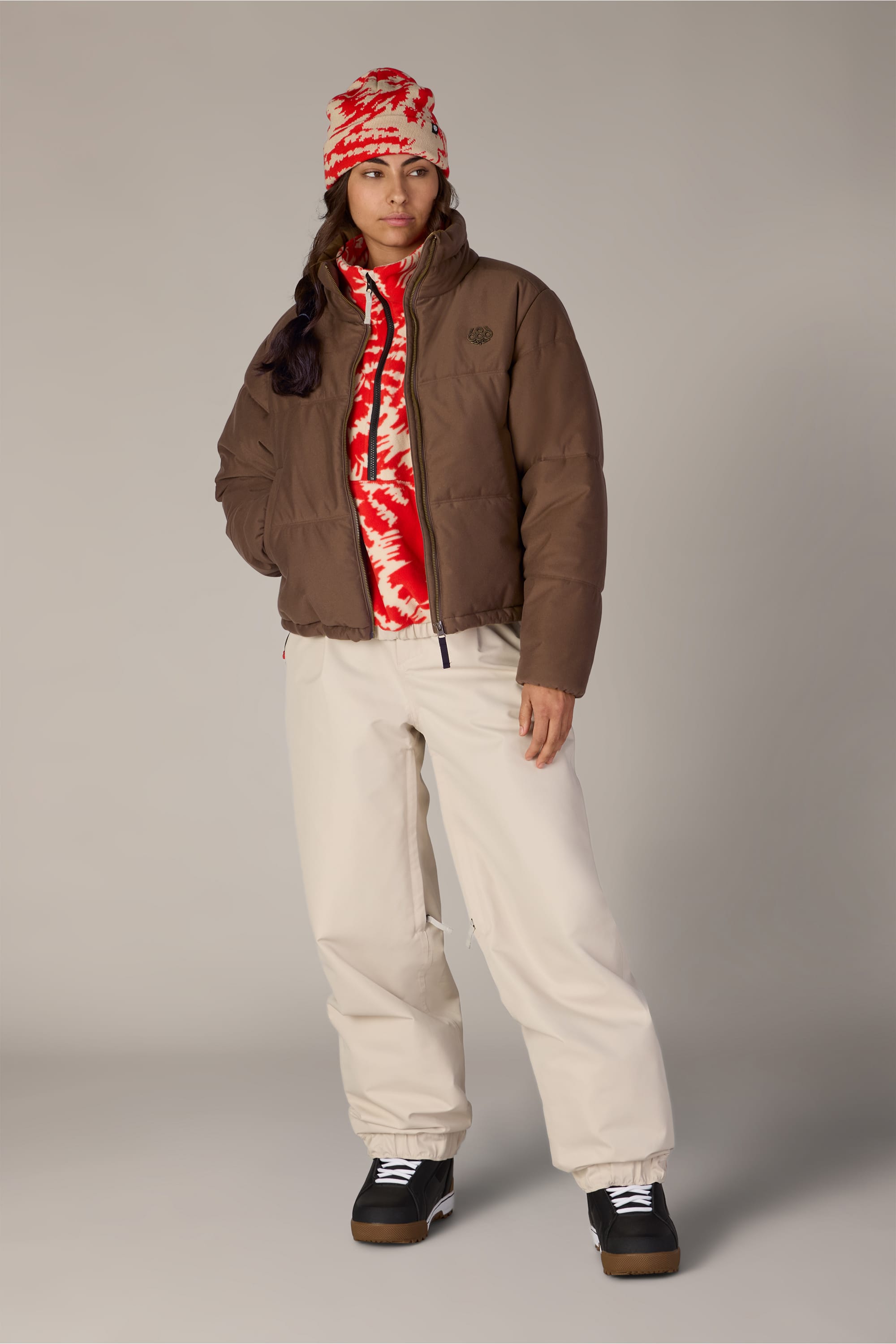 A person wearing the 686 Women’s Gracious Puffer Jacket in brown by 686, paired with a red-and-white patterned beanie, coordinating zip-up, cream pants, and black sneakers stands against a plain background with one hand in their pocket and a neutral expression.