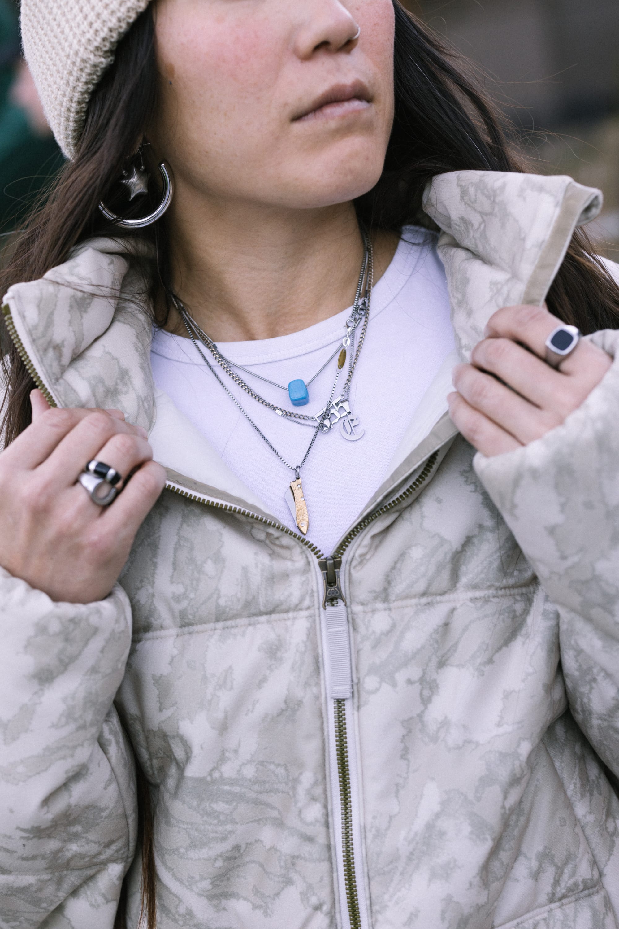 Wearing the 686 Women’s Gracious Puffer Jacket by 686, a person in layered necklaces, rings, and hoop earrings pulls up the jacket’s collar while looking away, pairing the look with a knit hat.