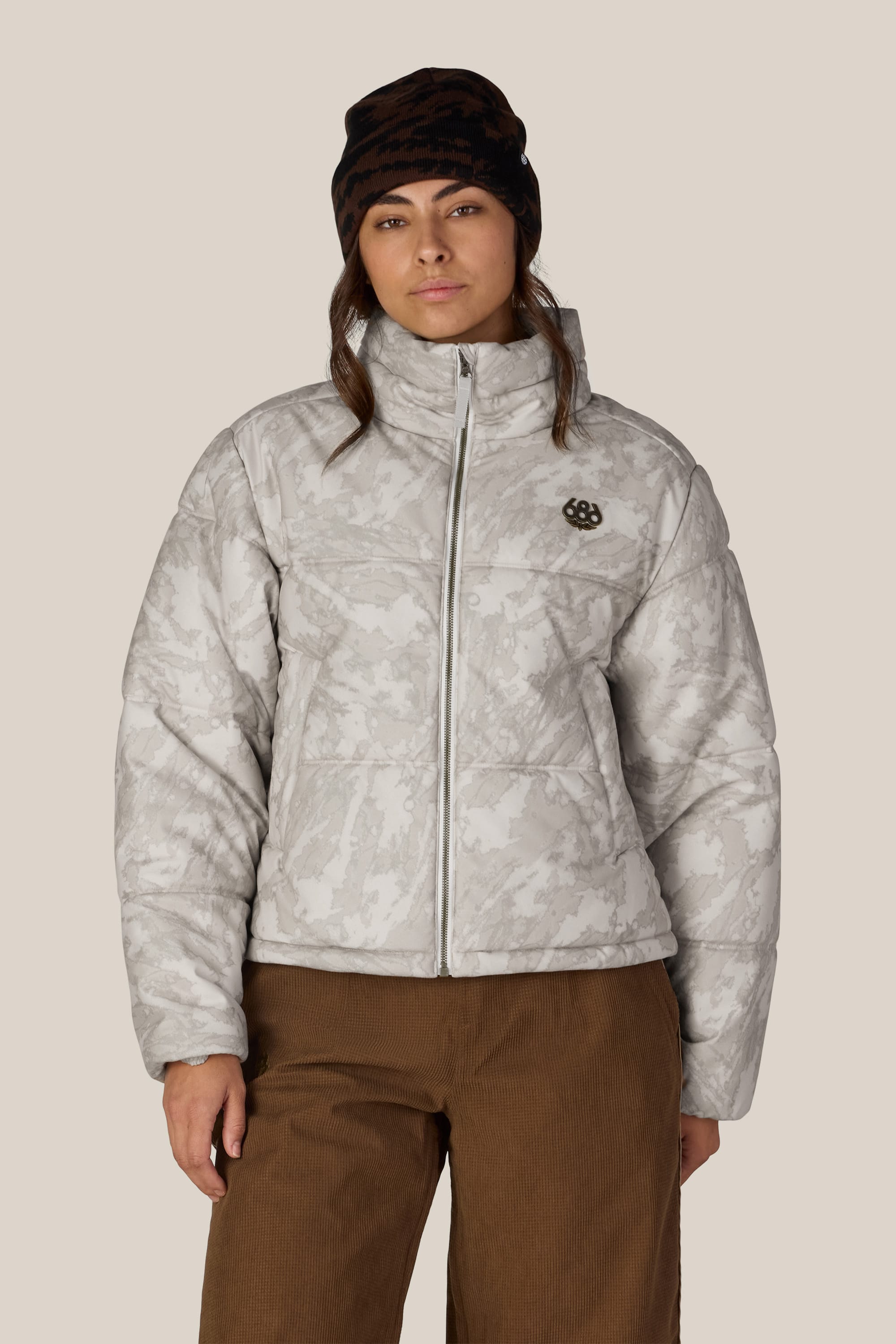A person wearing the 686 Women’s Gracious Puffer Jacket in light gray pattern, paired with brown corduroy pants and a black beanie, stands against a plain beige background facing forward with a neutral expression.