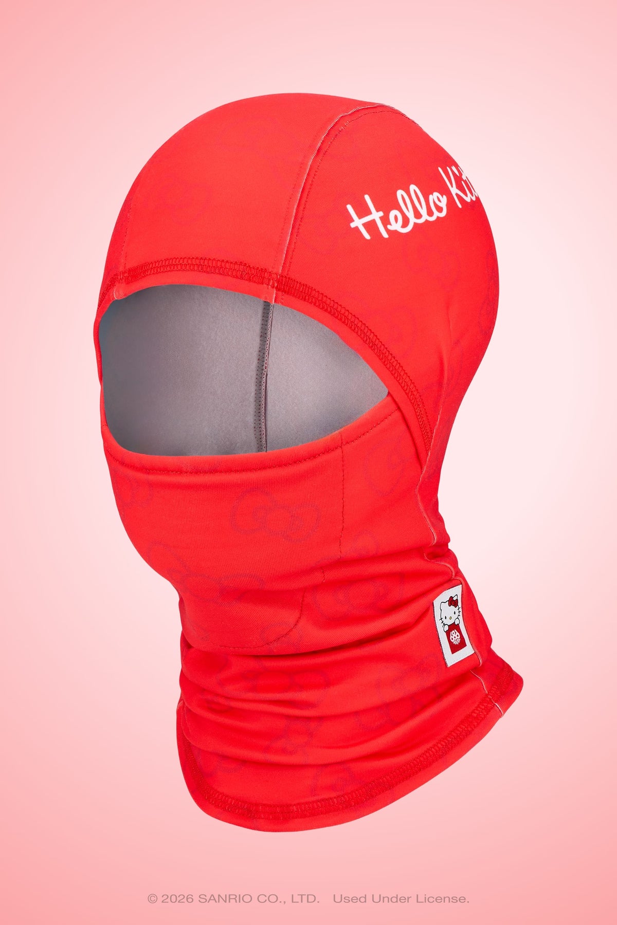 A red 686 Hurra Hinged Balaclava featuring Hello Kitty written on the forehead and a small Hello Kitty patch on the chin, displayed on a pink gradient background.