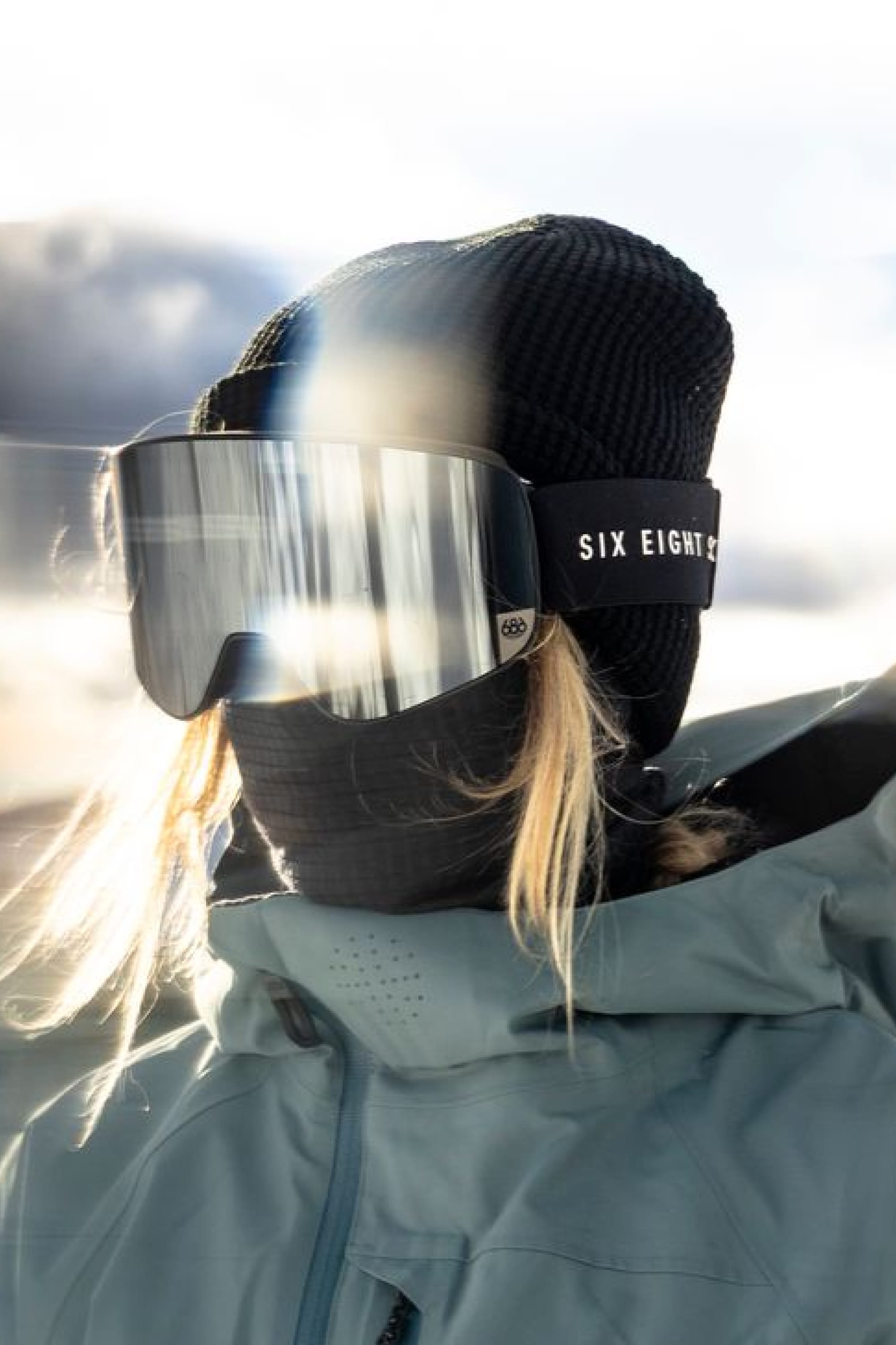A person wearing the 686 Mission Grid™ Balaclava and ski goggles, dressed in a light blue winter jacket, stands outdoors with sunlight reflecting off the goggles.