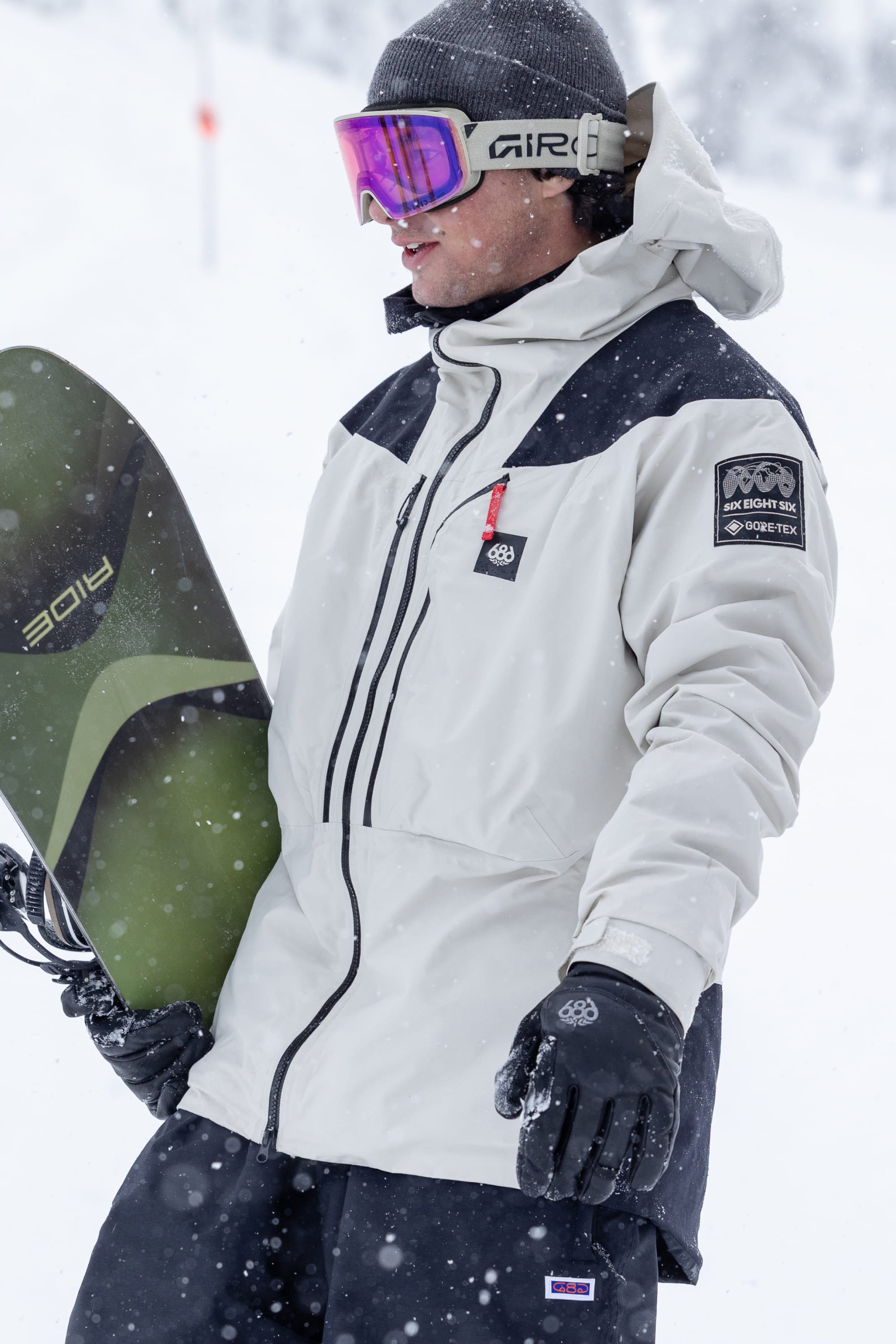 A person in a white winter jacket, wearing the 686 Men’s GORE-TEX® Stronghold Leather Glove, a beanie, and ski goggles holds a snowboard in a snowy outdoor setting with snow visibly falling.