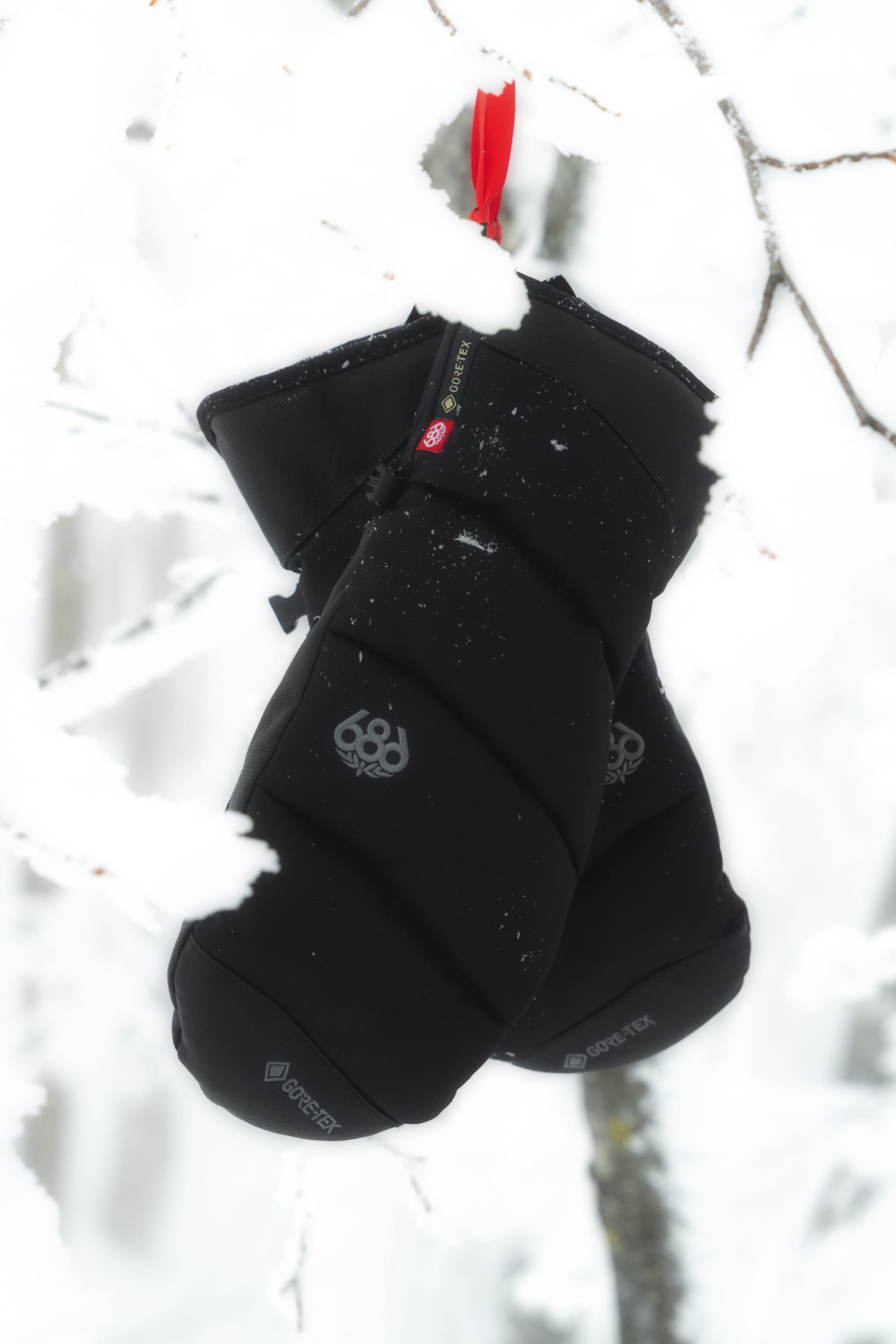 A pair of 686 Men’s GORE-TEX® Buffer Insulated Mitts, in black with a red loop, hang from a snow-covered tree branch against a white snowy background.