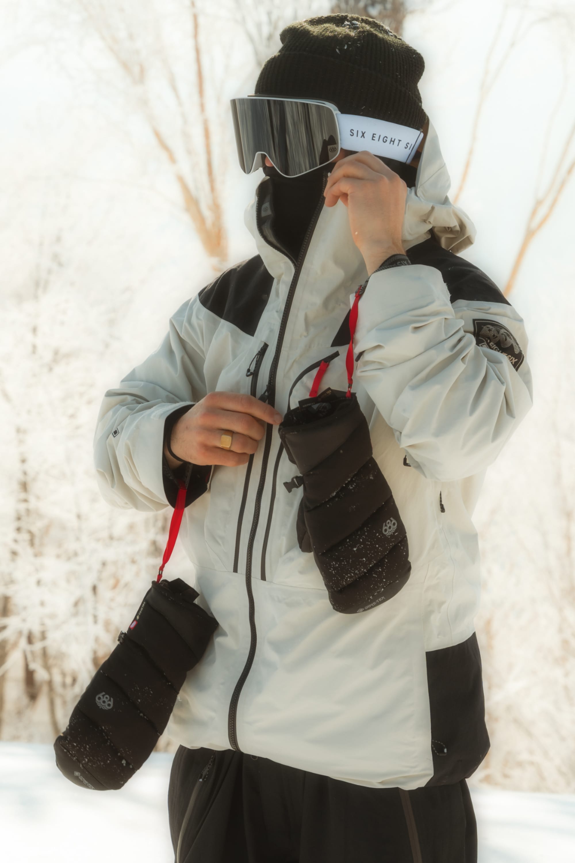 A person in a white winter jacket, ski goggles, and beanie stands outdoors in the snow, holding up a 686 Men’s GORE-TEX® Buffer Insulated Mitt near their face. Two cylindrical black pouches hang from red straps around their neck.